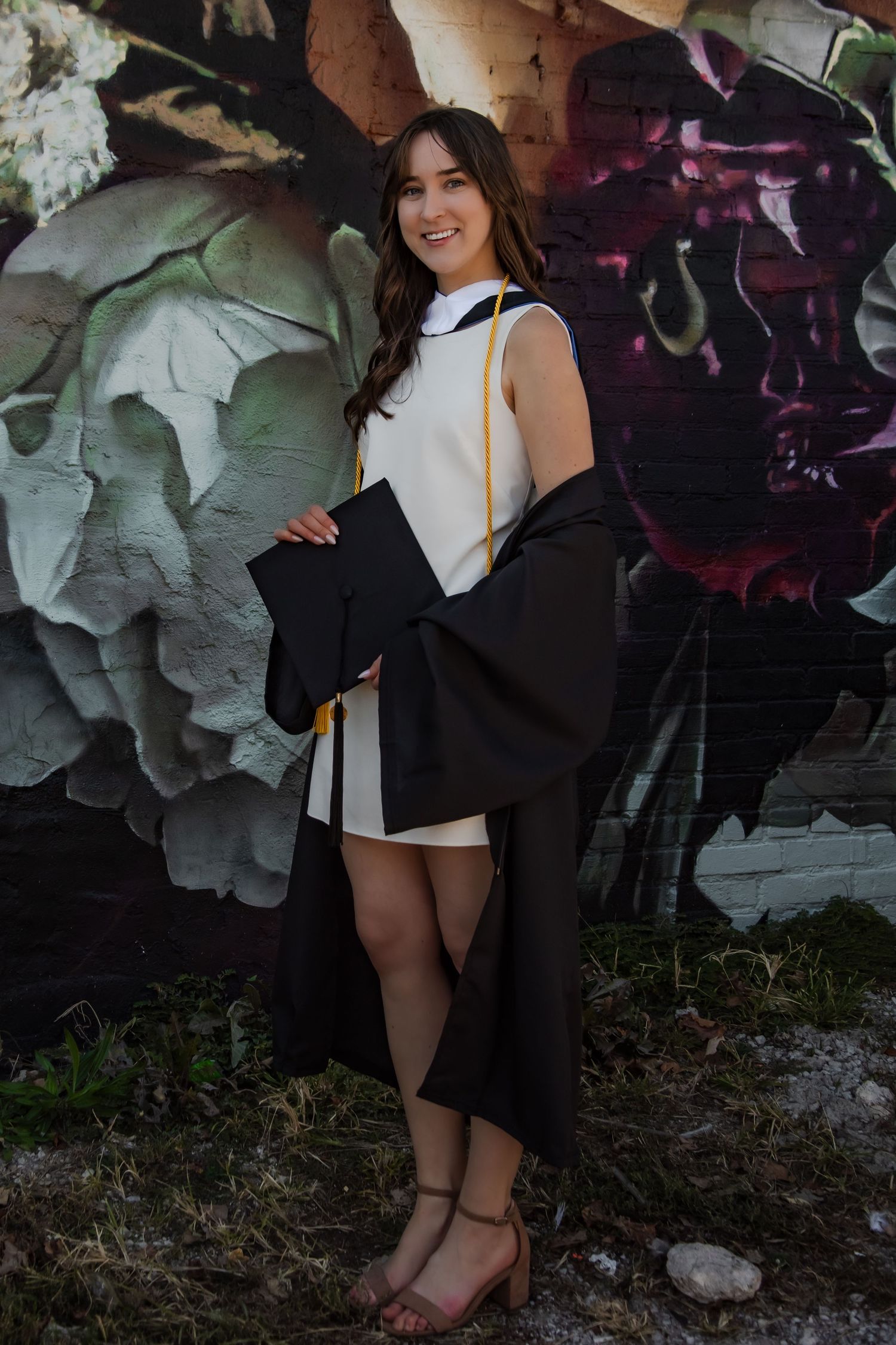 Graduate in white dress and black gown poses against artistic floral wall mural.
