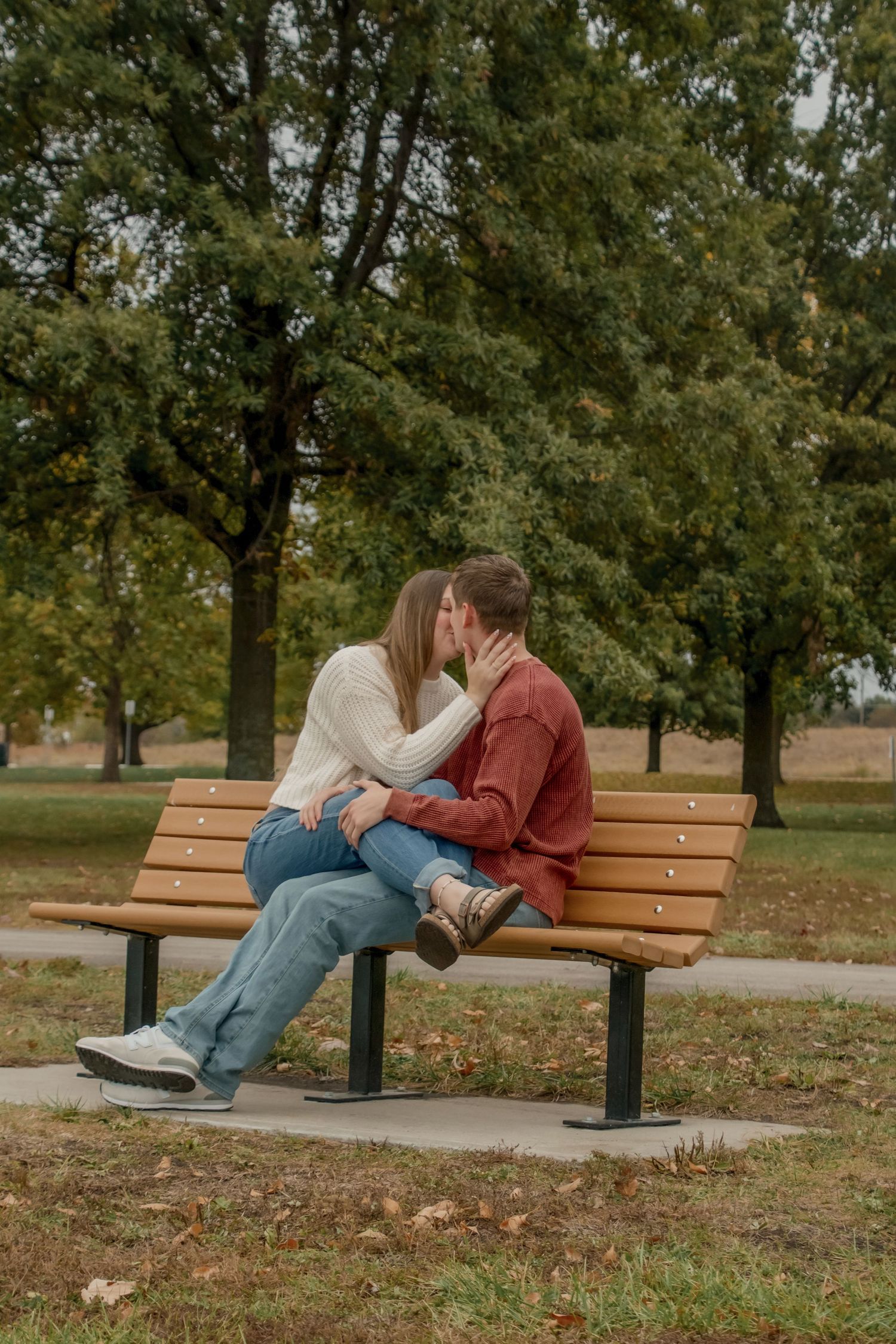A couple relaxes together on a wooden park bench surrounded by autumn trees and fallen leaves.