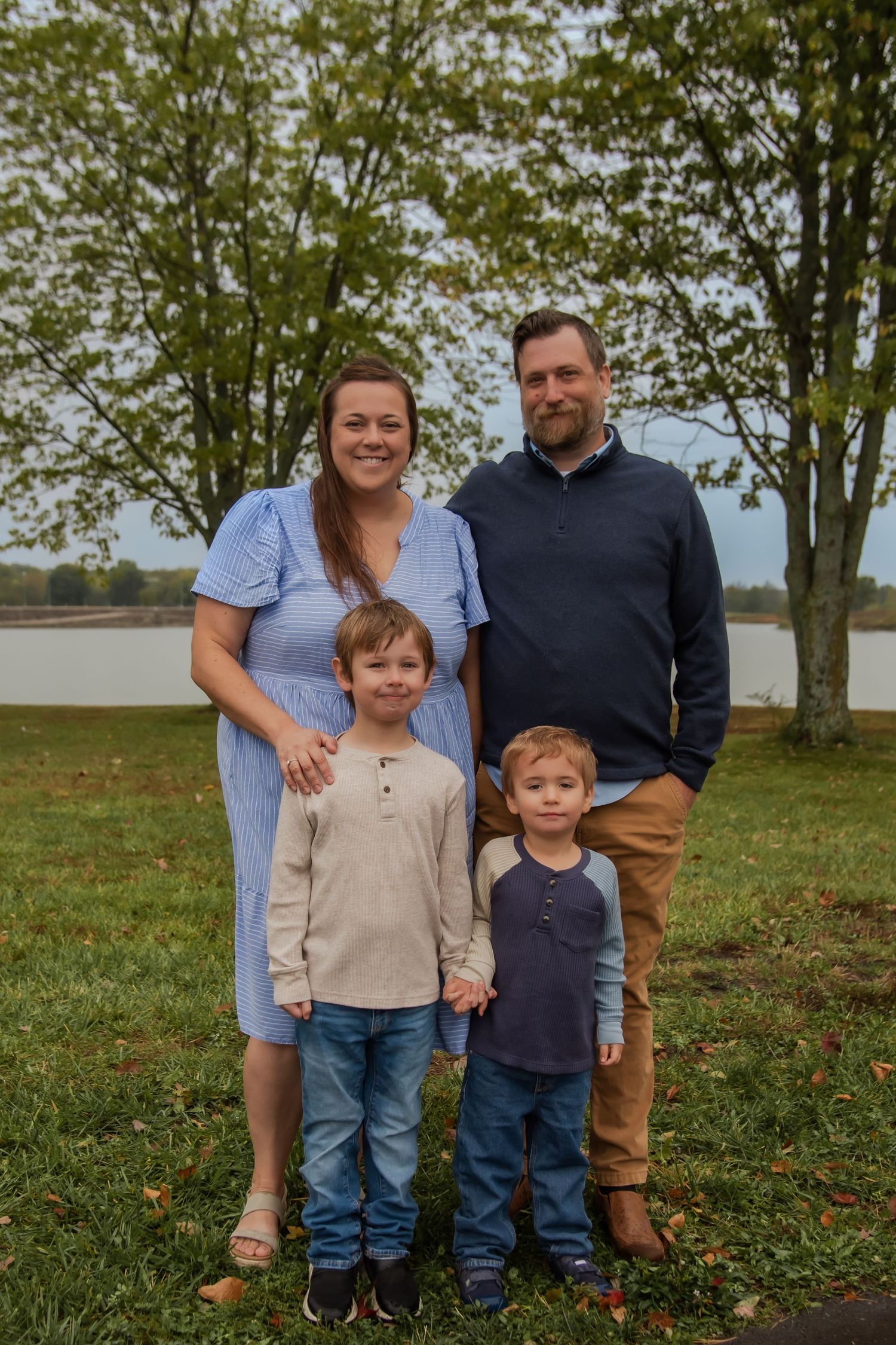 A family of four poses together by a lake on an autumn day with trees in the background.