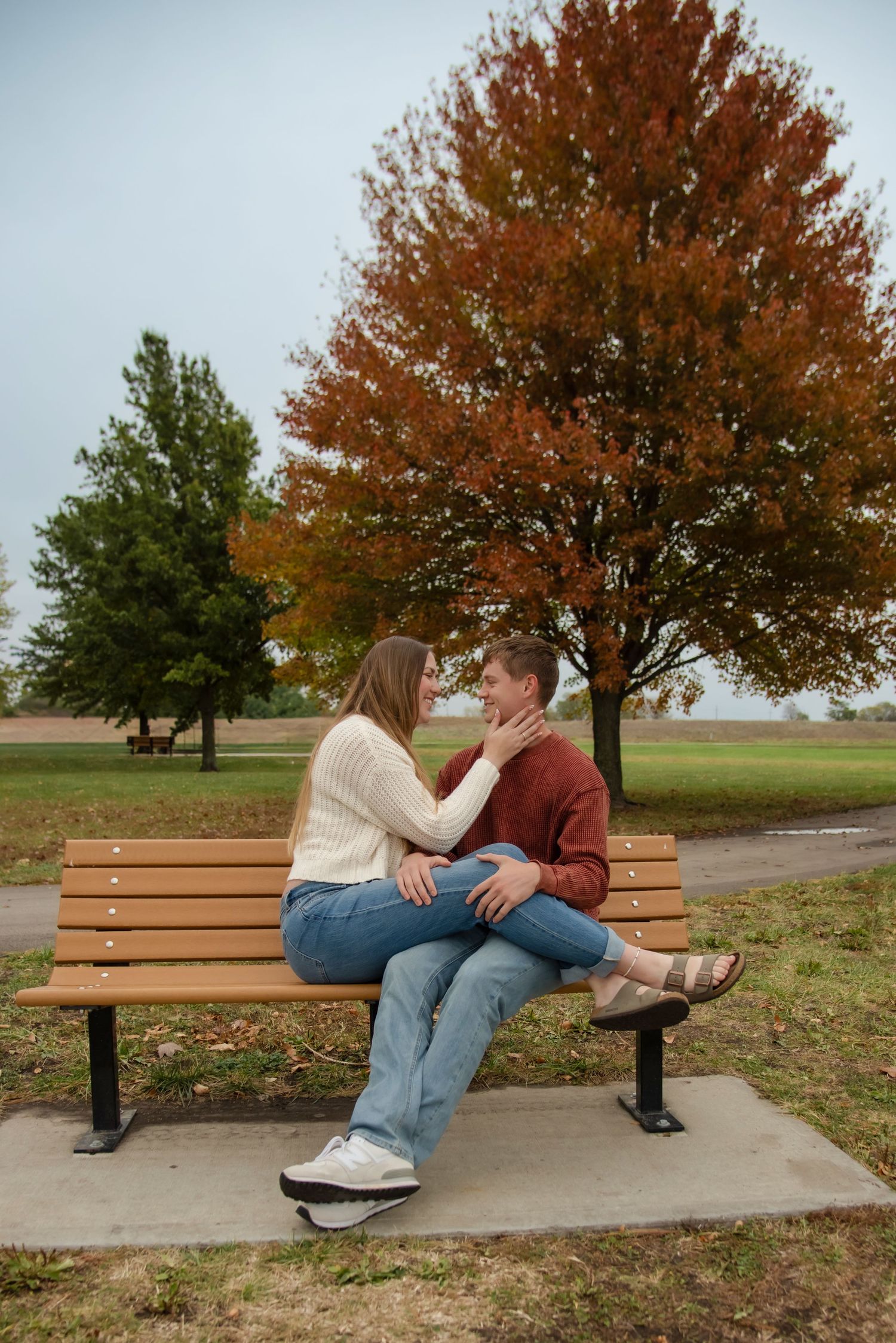 Two people sit together on a wooden park bench under autumn trees with orange leaves on a cloudy day.