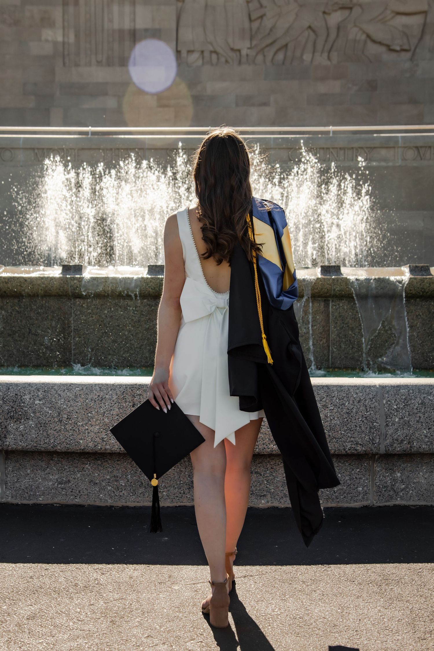 Graduate in cap and gown poses near fountain during outdoor photo session.