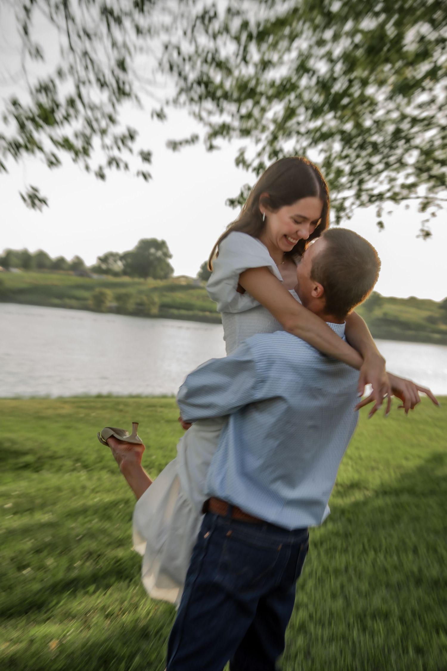A couple shares a playful embrace while spinning in circles on green grass near a lake.