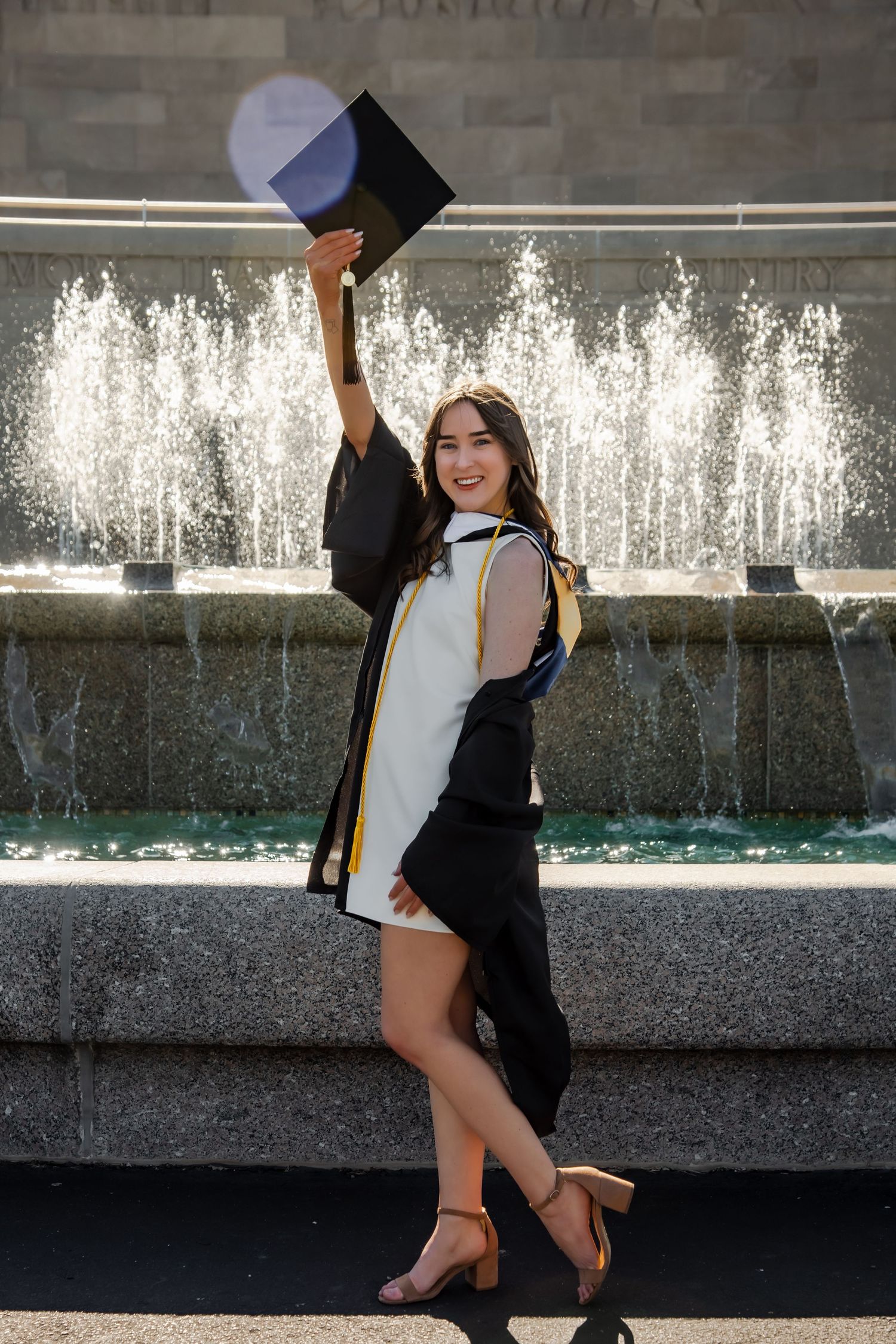 Graduate in black gown and white dress poses playfully near outdoor fountain.