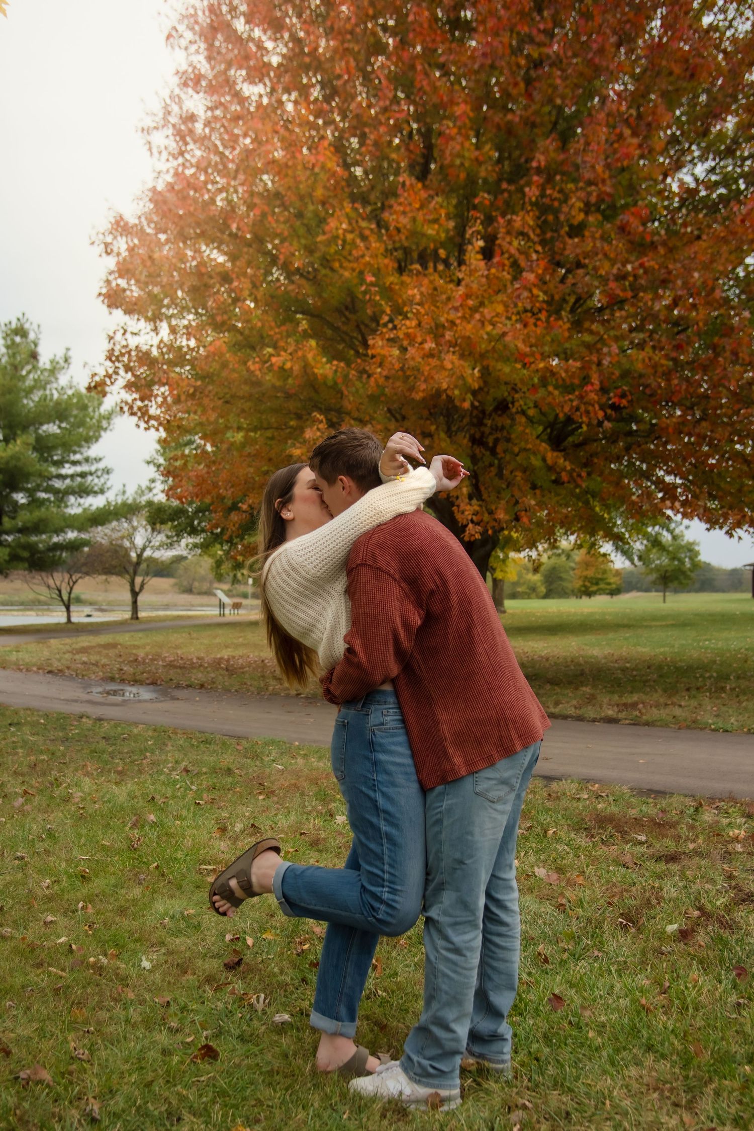 A couple shares a playful moment together on an autumn path surrounded by trees with vibrant orange foliage.