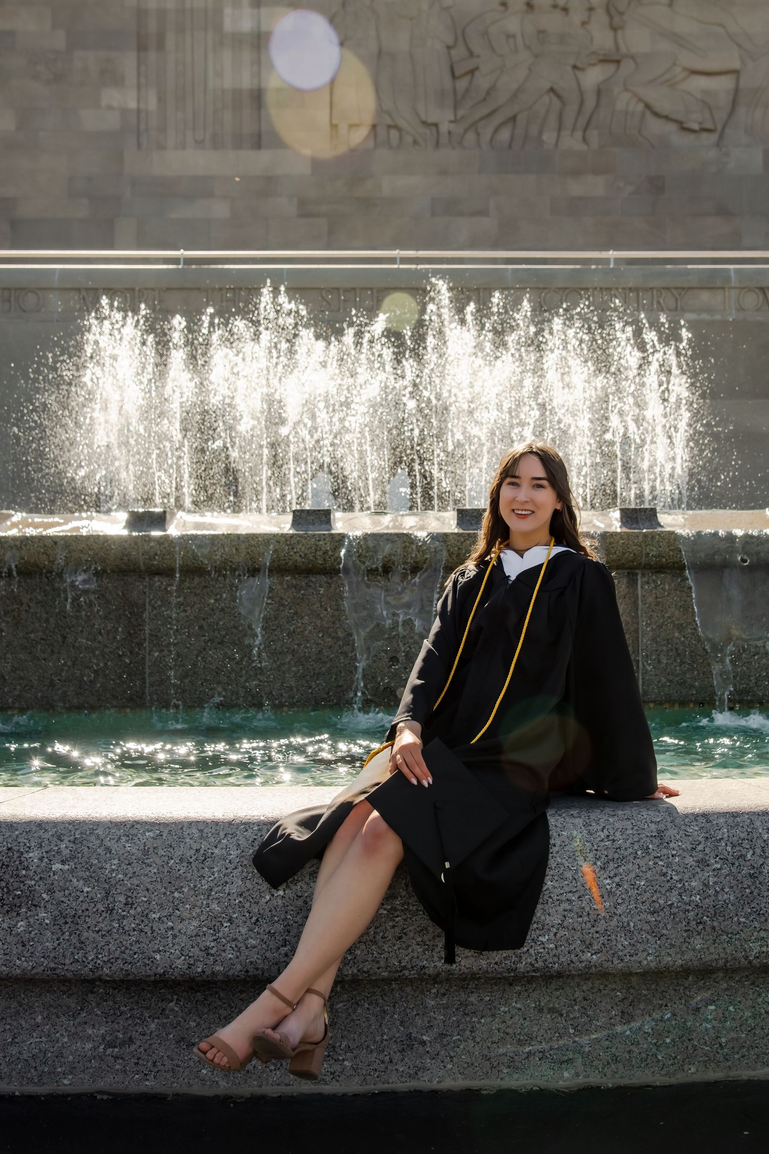 Graduate in cap and gown sitting by fountain with water sprays in background.