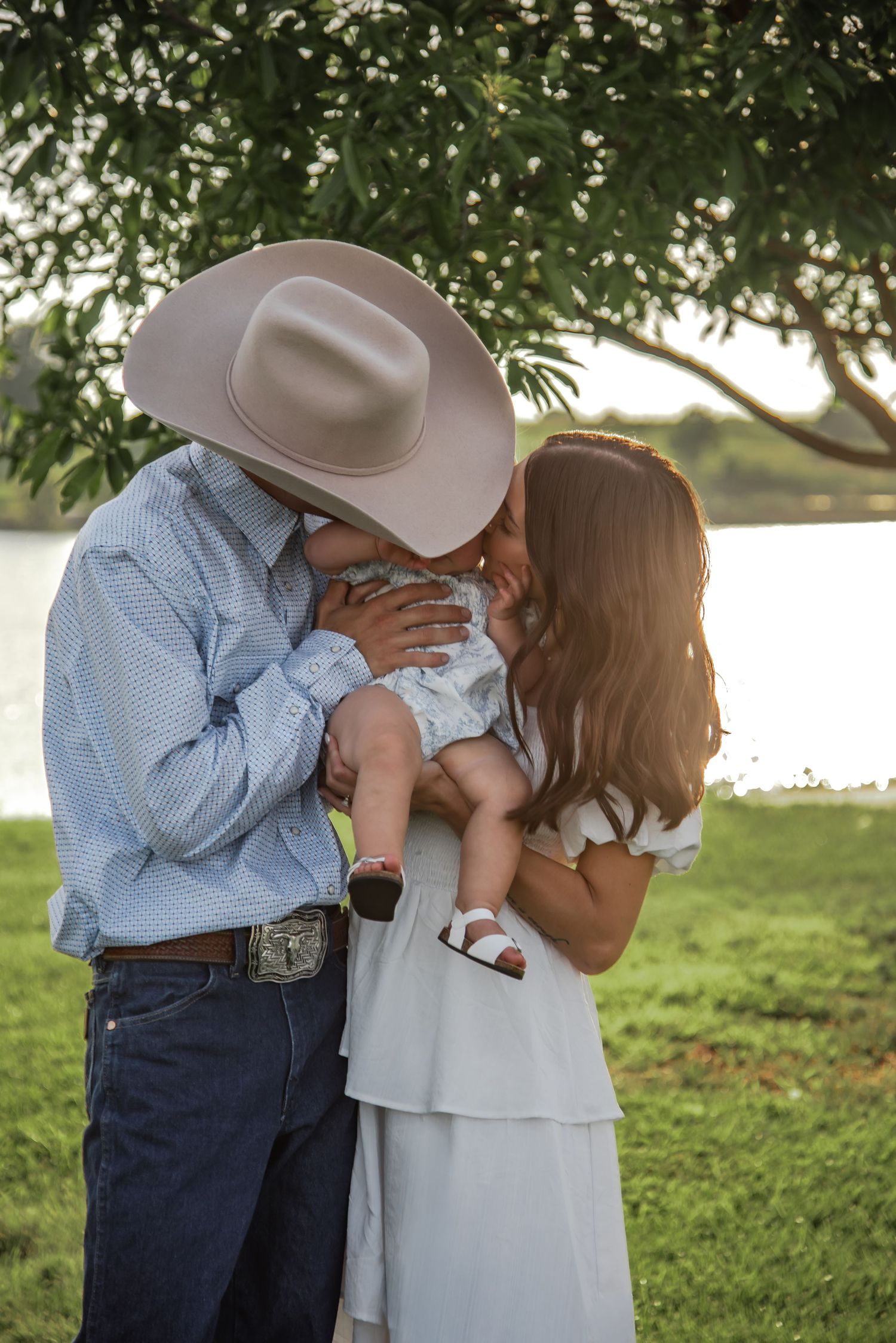 A couple in western attire shares a romantic kiss outdoors under trees in a parklike setting.