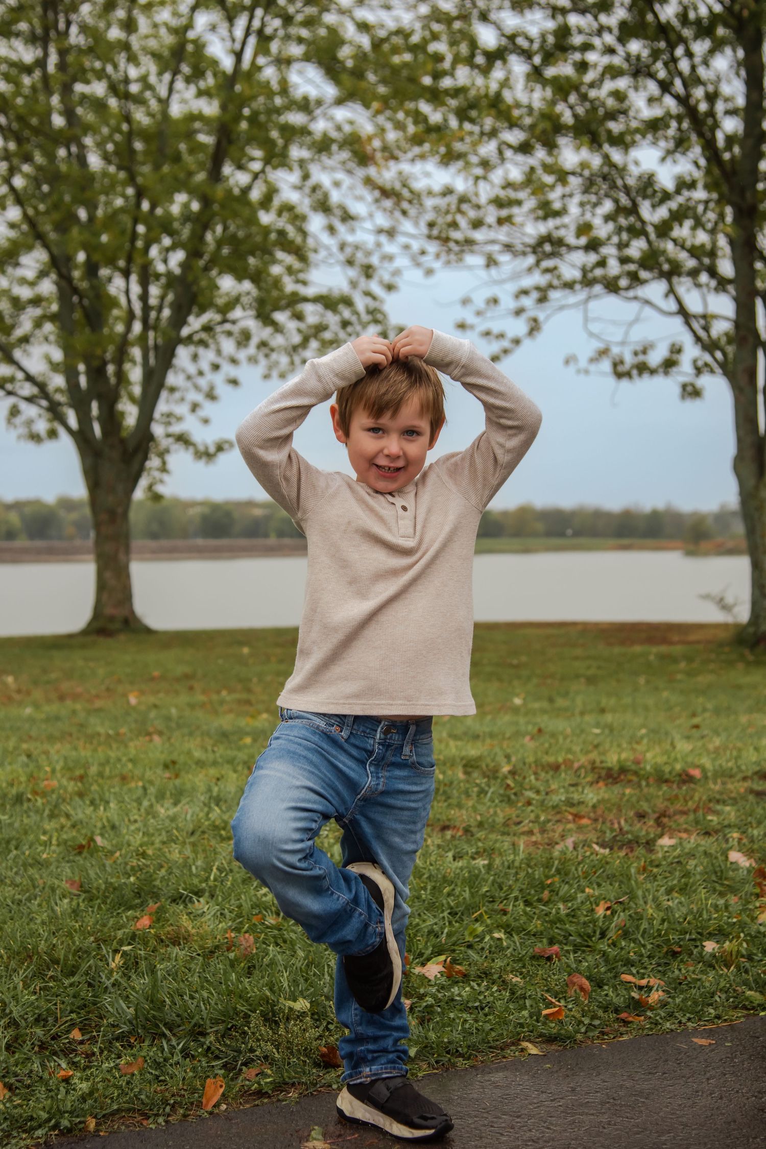 A playful figure in a beige sweater and jeans dances and jumps on a path surrounded by autumn trees.