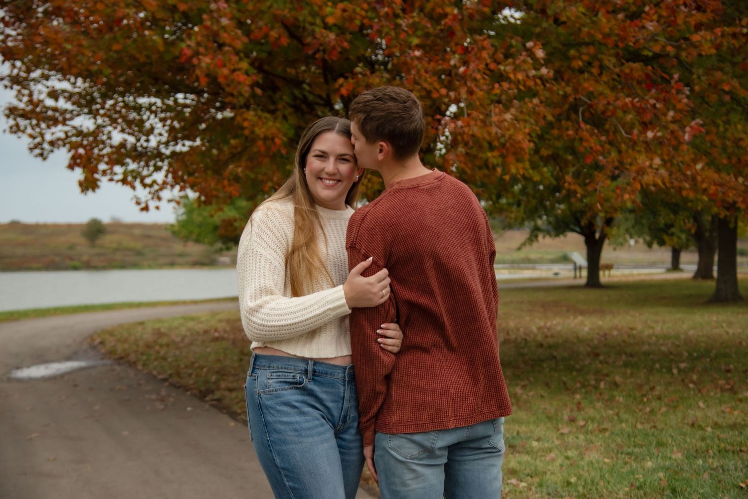 A couple embraces while walking through a park with autumn trees displaying vibrant orange and red foliage.