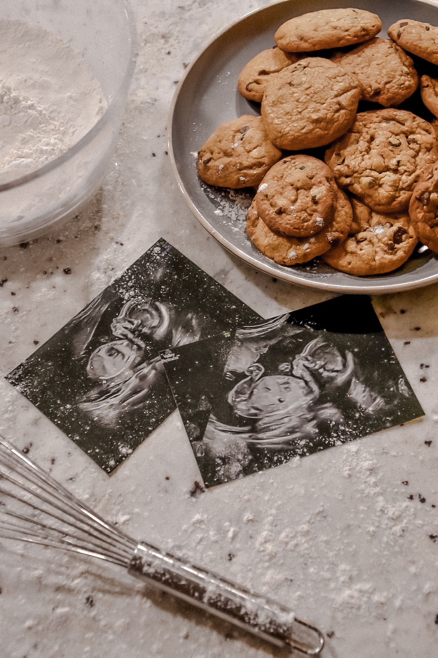 Fresh baked chocolate chip cookies on a plate with scattered flour and vintage recipe photos on a concrete surface.