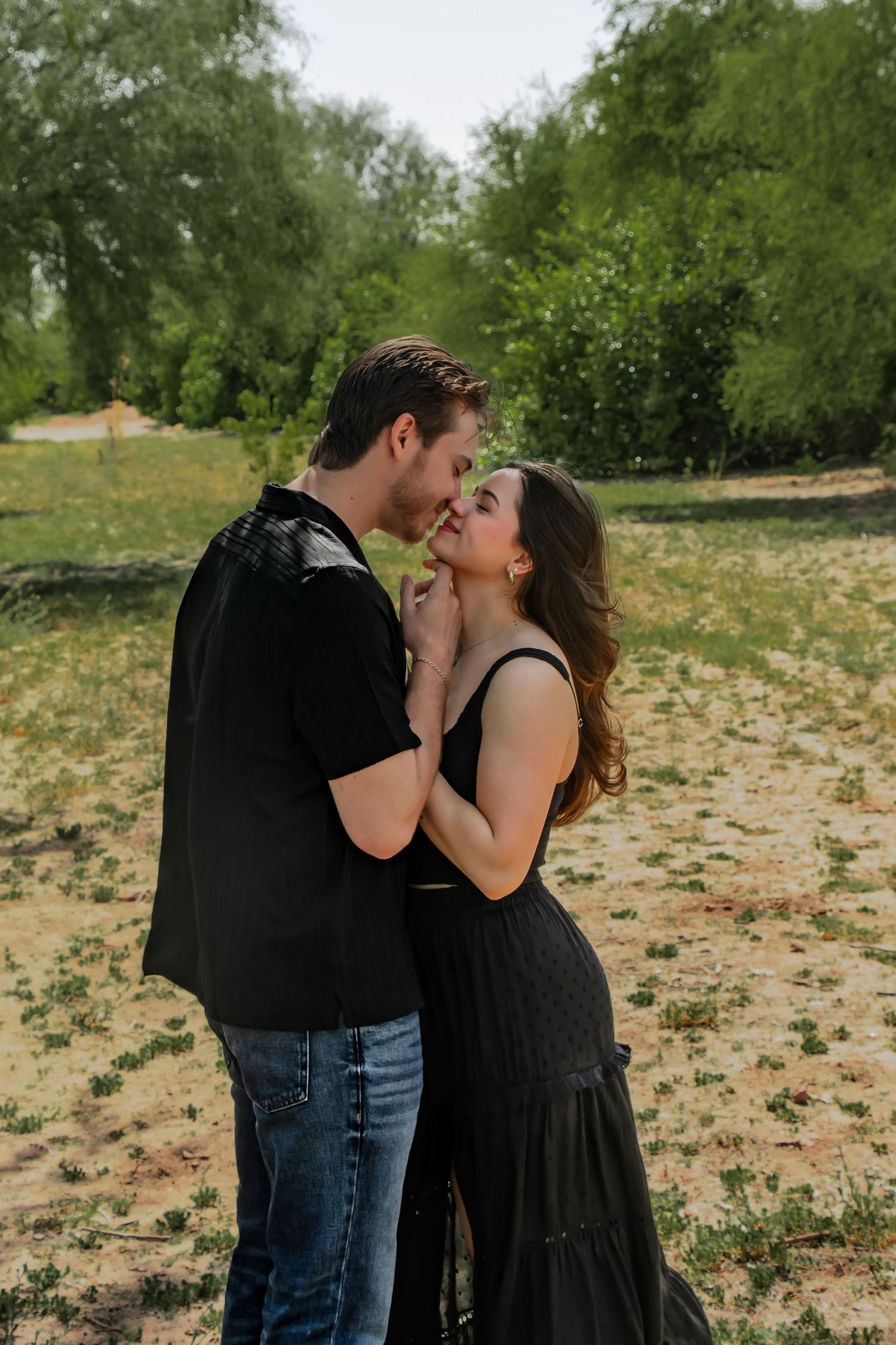 A couple wearing black clothing share a romantic kiss in a sunny field with green trees in the background.