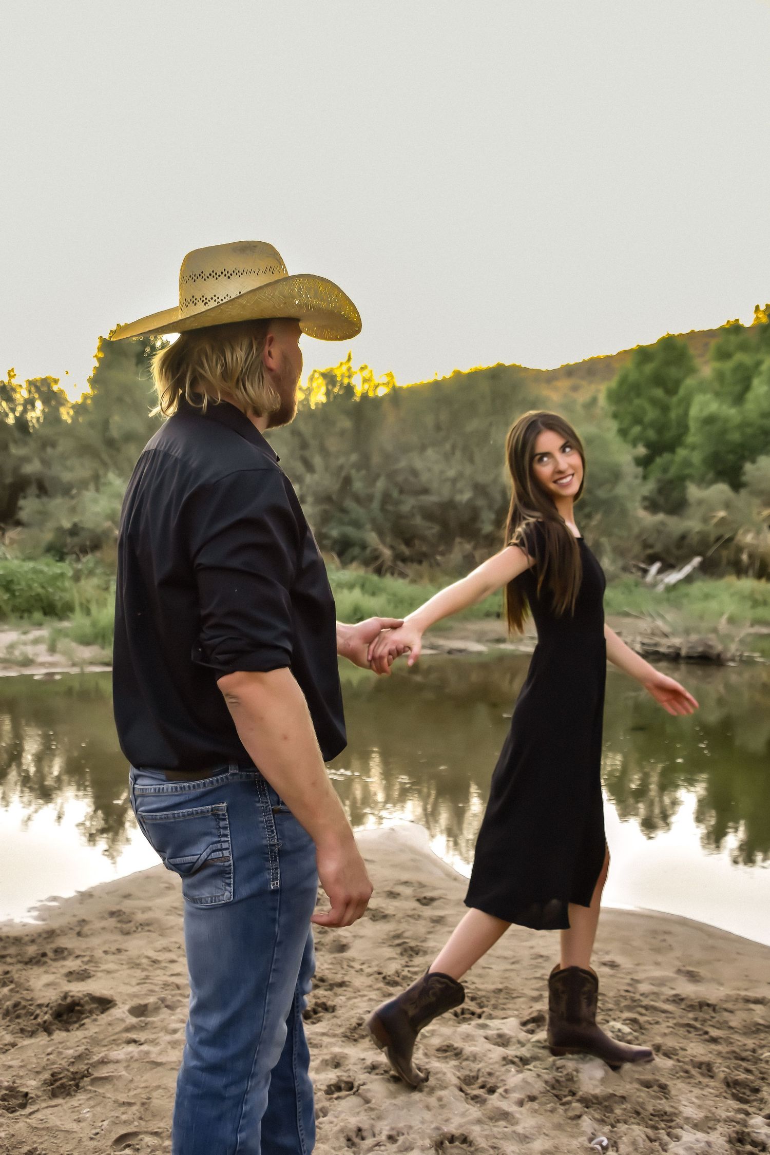 Couple in western wear shares playful dance moves on lakeside beach at sunset.