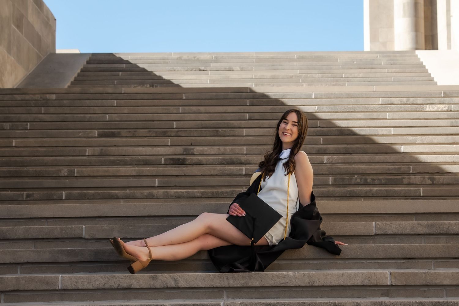A person in casual attire sits on concrete steps on a sunny day, with a backpack beside them.