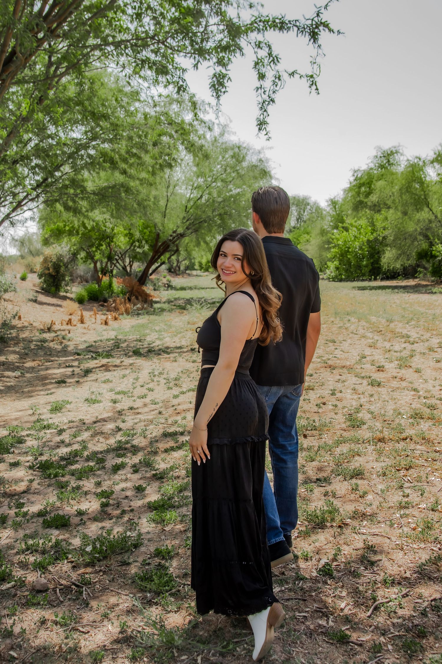 A romantic couple walks together on a desert path surrounded by green trees and natural terrain.