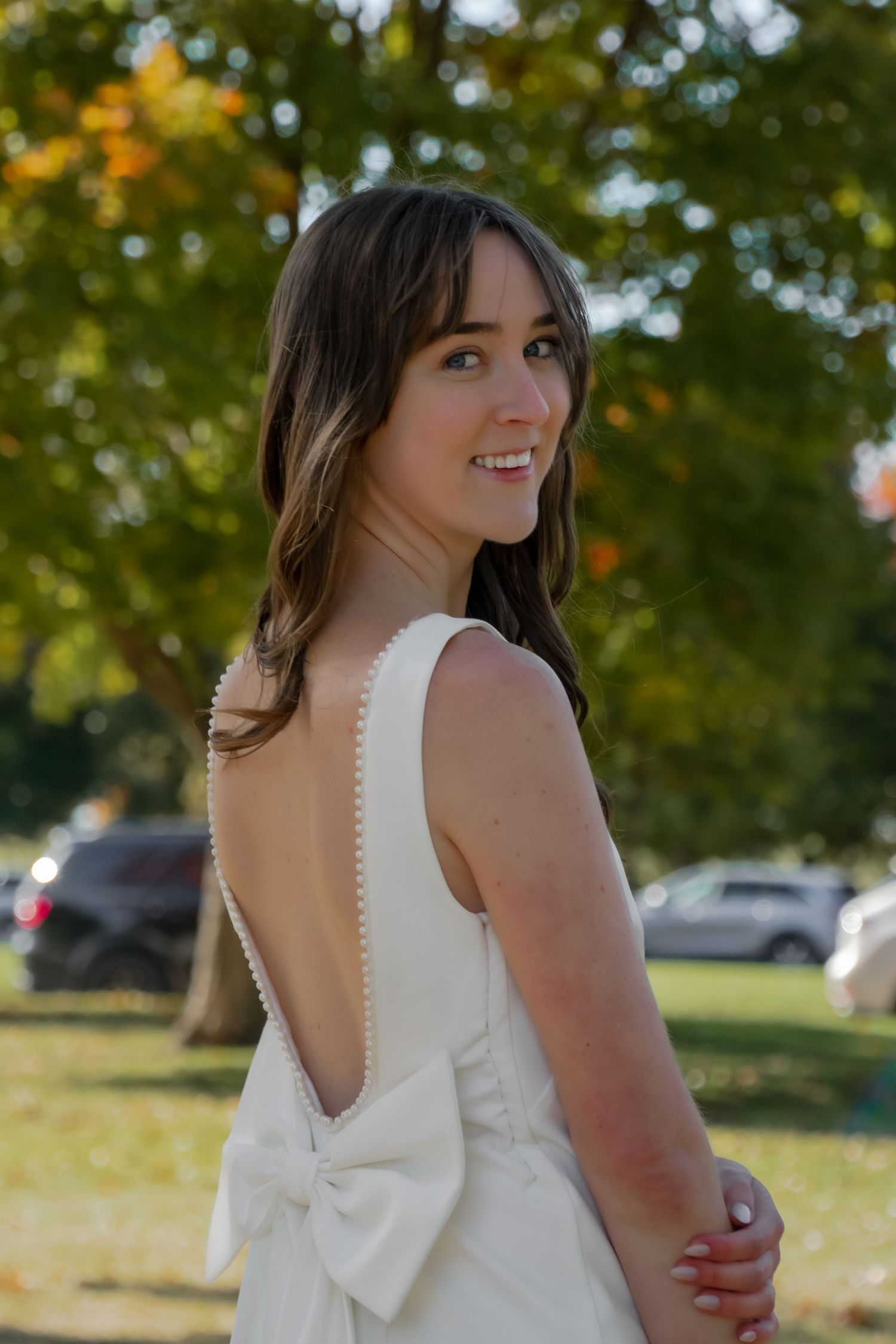 Graduate in white dress with bow detail poses outdoors during autumn season.