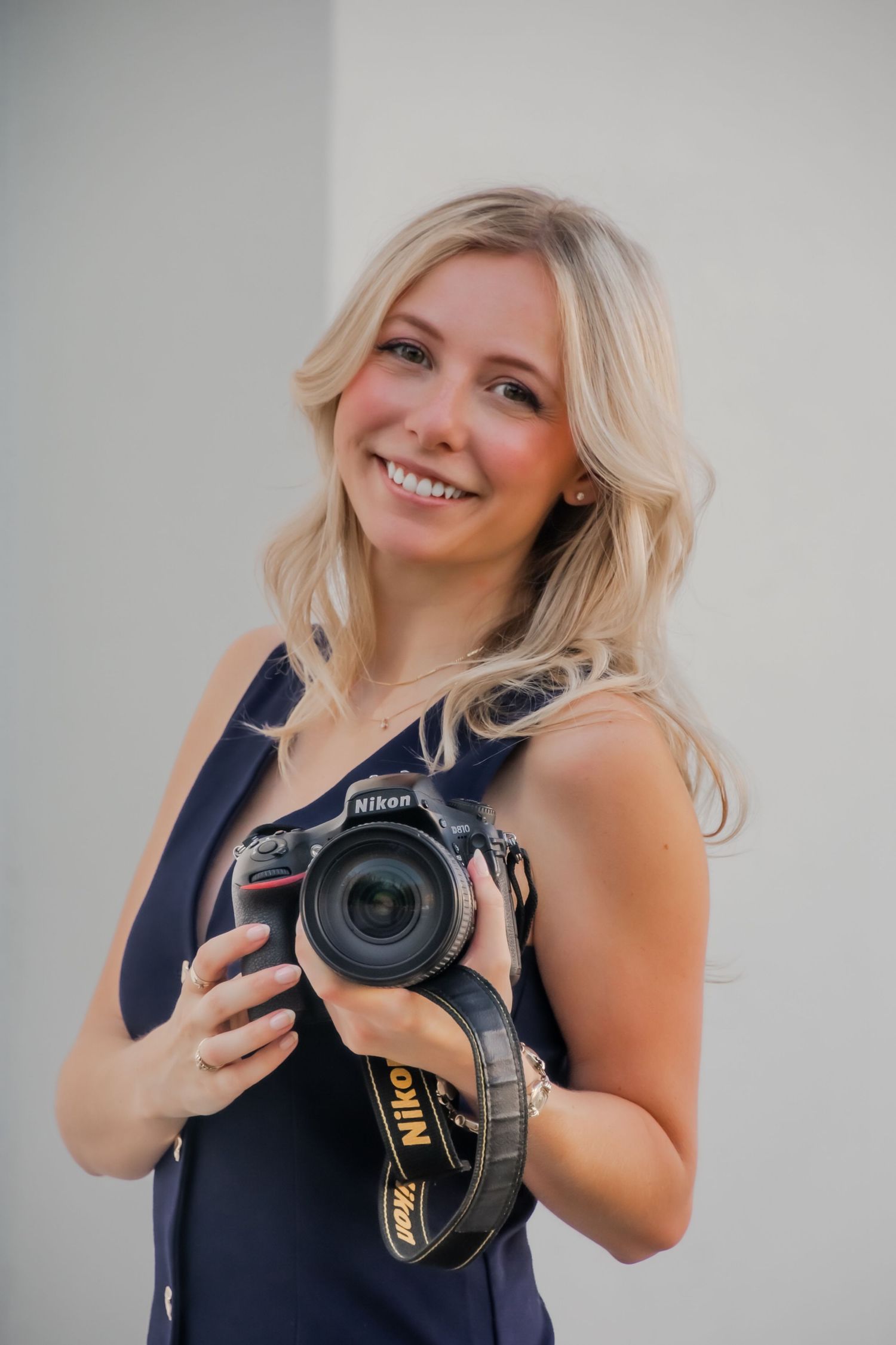 Professional photographer in navy top smiling and posing with a Nikon DSLR camera.