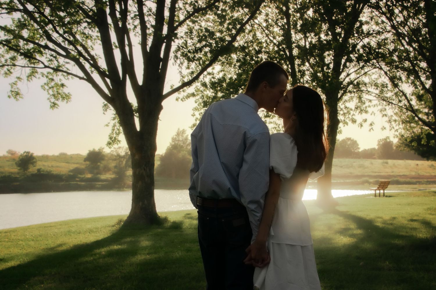 Silhouetted figures embrace beneath towering trees along a peaceful lakeside at golden hour.