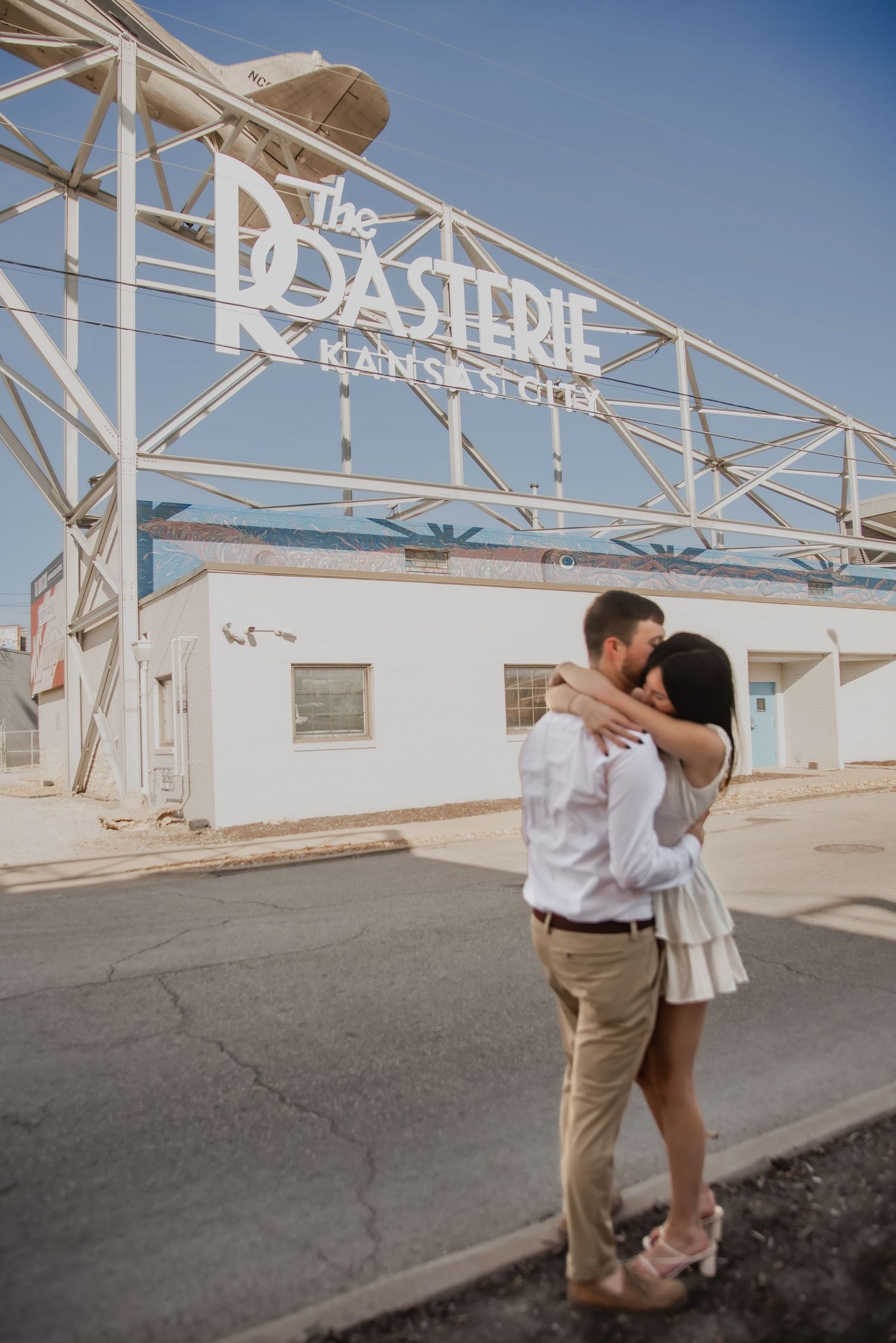 A couple shares a romantic moment outside a white building with 'Pastene' signage on a sunny day.