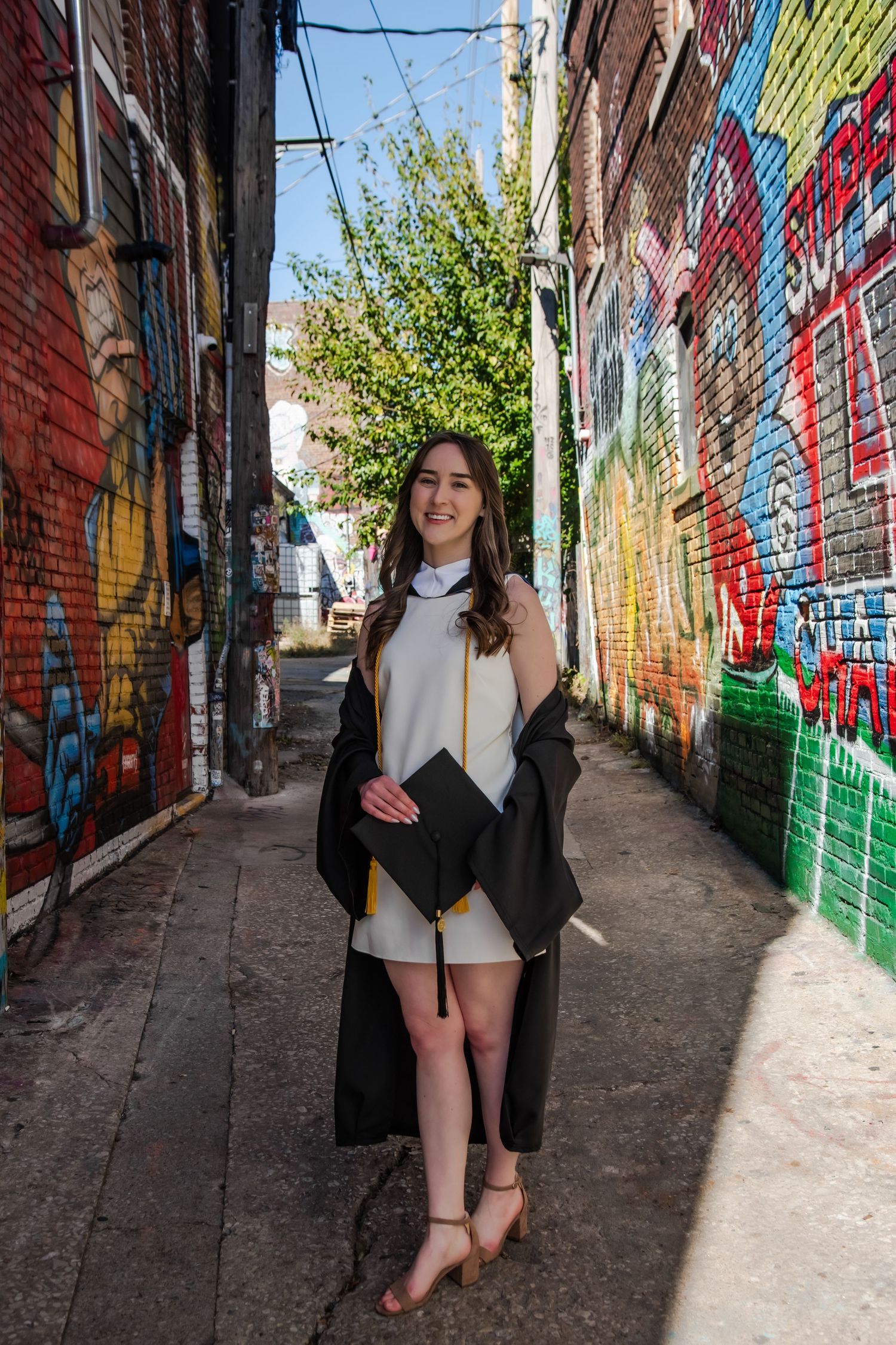 Graduate in academic dress poses in colorful graffiti alley with brick walls.