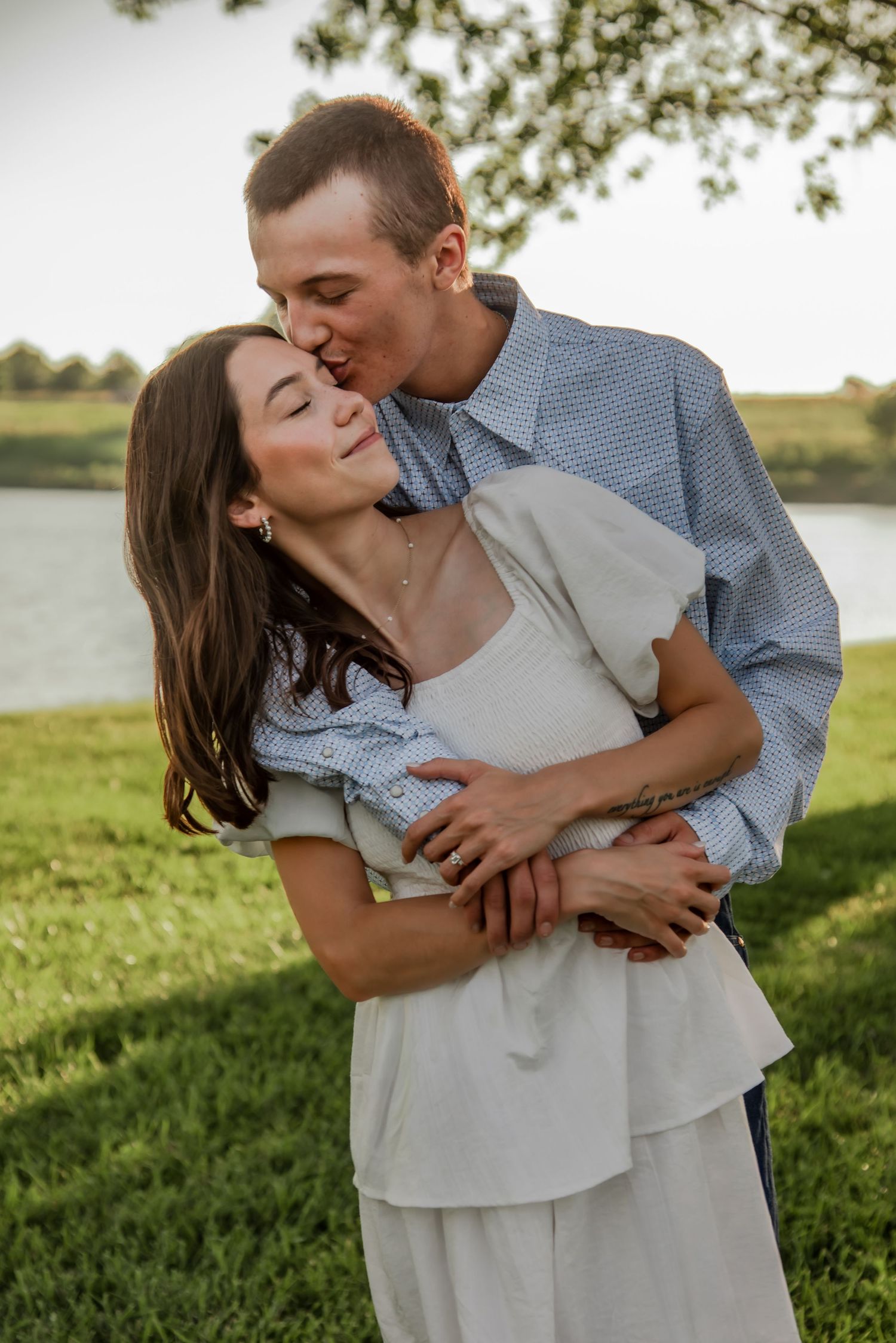 A couple shares an intimate moment on a sunny day by a lake with lush green grass in the foreground.