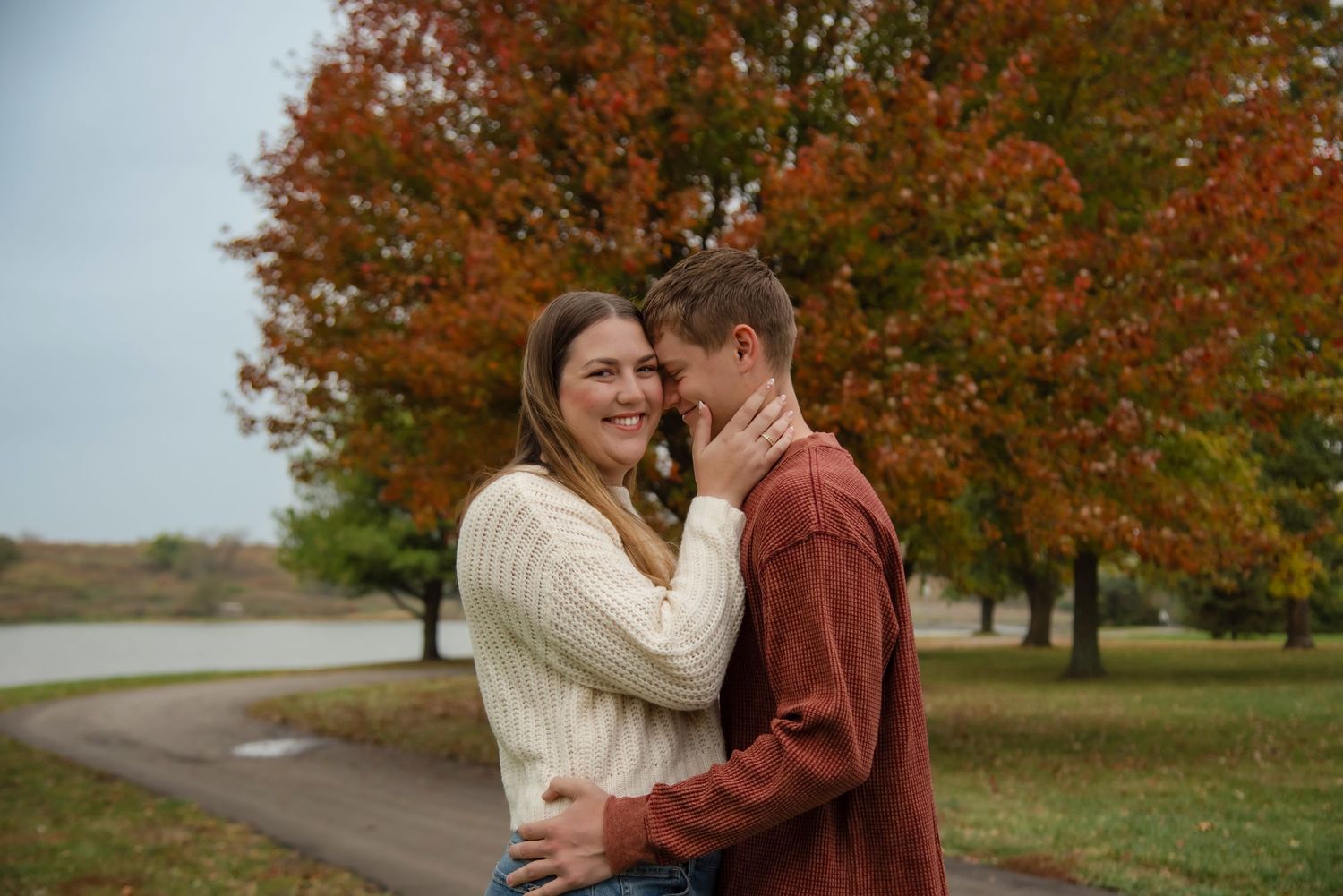 A couple embraces outdoors during autumn with vibrant orange fall foliage in the background.