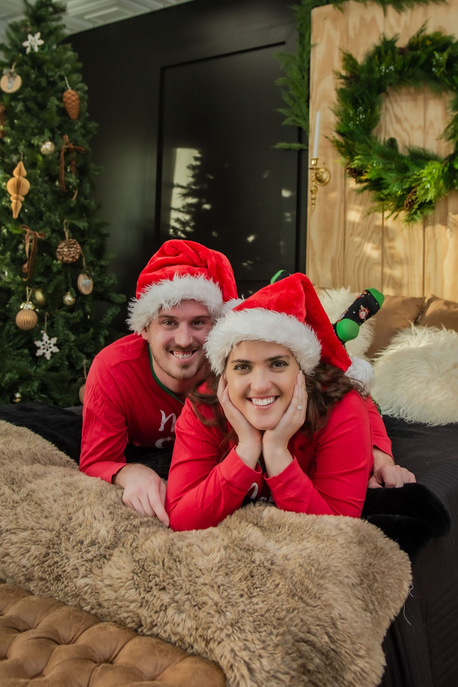 Couple in red Santa hats and outfits lying on a brown fur rug with Christmas decorations in the background.