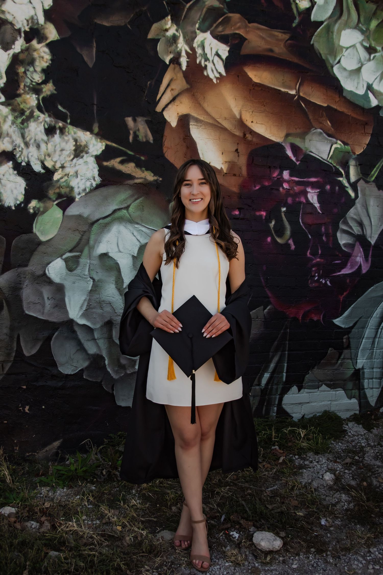 Graduate in graduation attire standing in front of artistic floral wall mural.