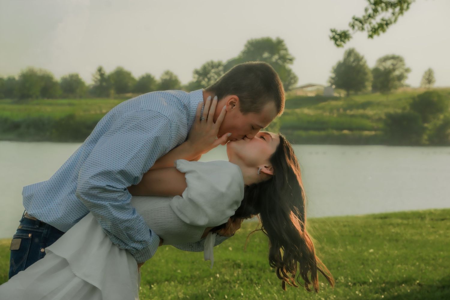 A person dips someone in a romantic kiss pose outdoors by a scenic lake on a sunny afternoon.