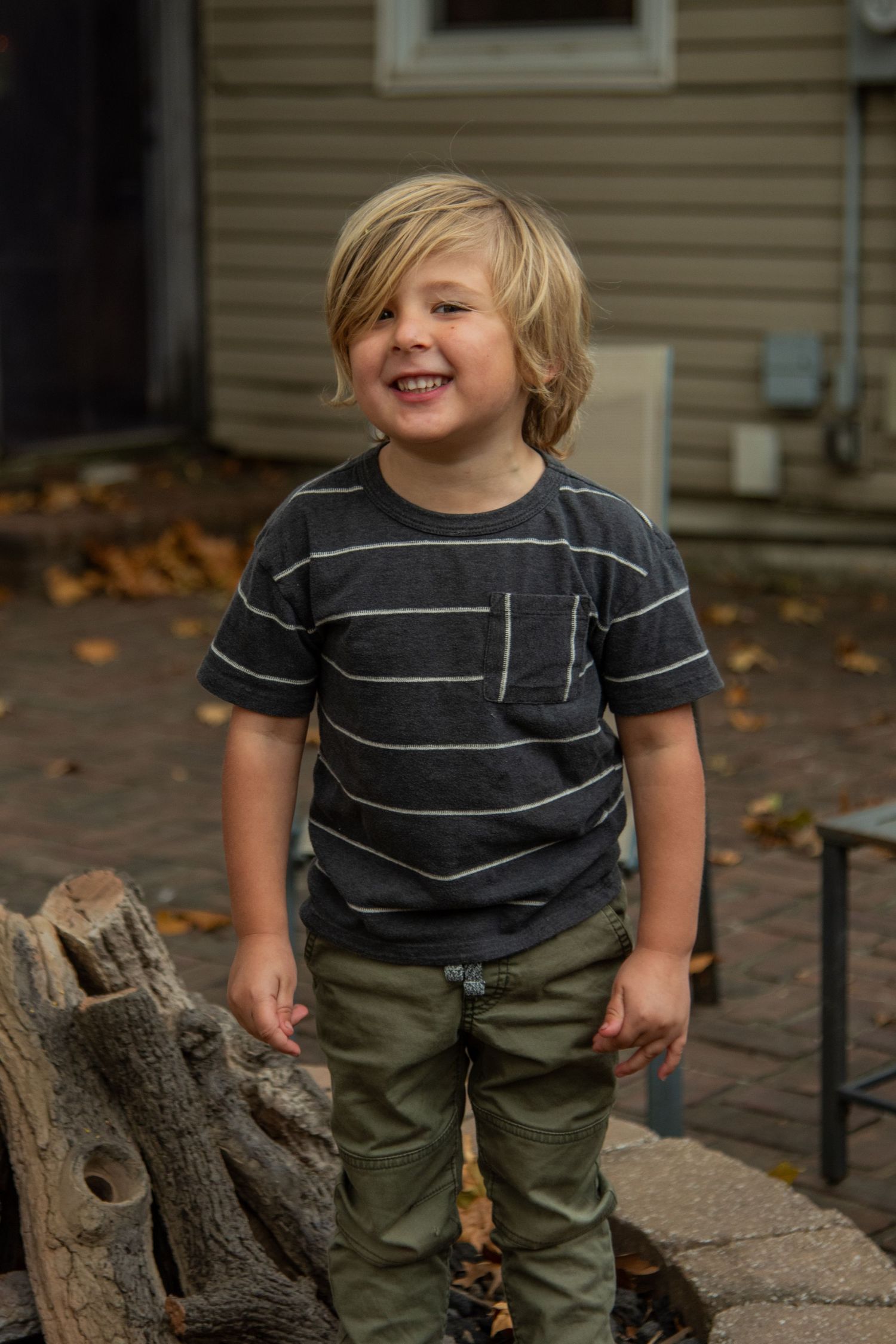 A young child in a striped shirt and olive pants smiles while standing outdoors.