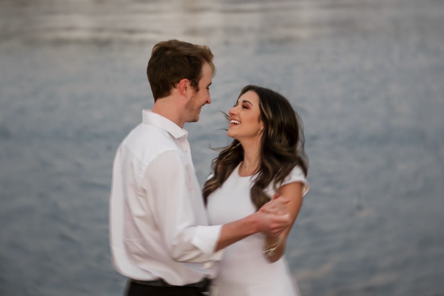 A romantic couple in formal attire shares an intimate moment by the water during a sunset photoshoot.