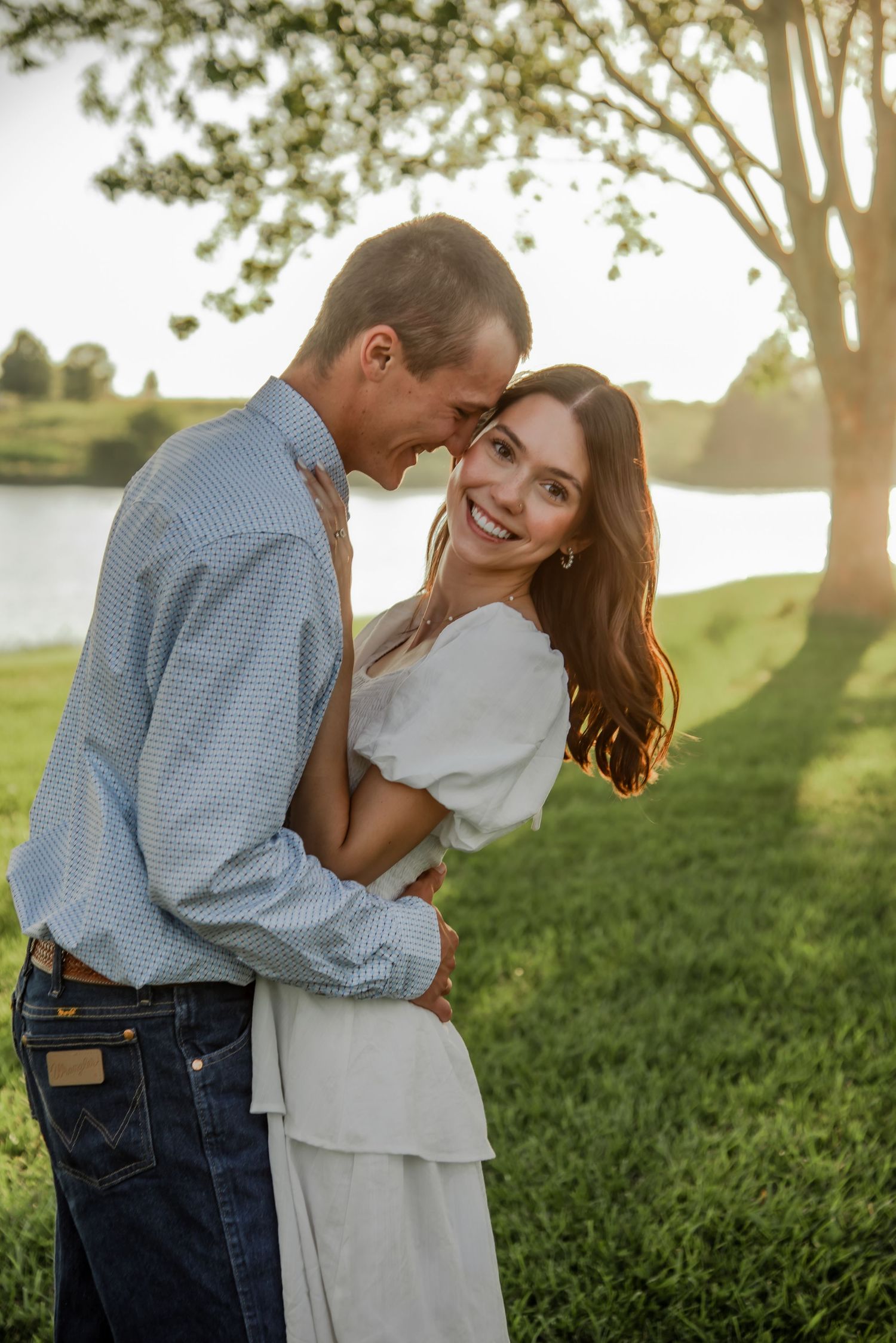 Romantic couple embraces in a sunlit field with trees during golden hour.