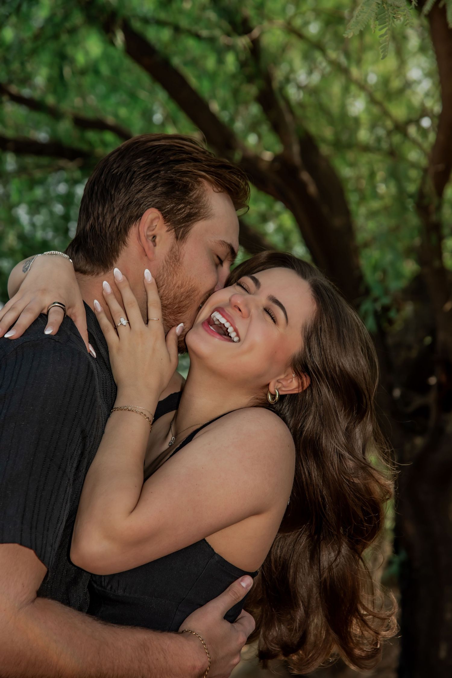 A romantic outdoor photoshoot captures a joyful embrace between a couple laughing together under green trees.