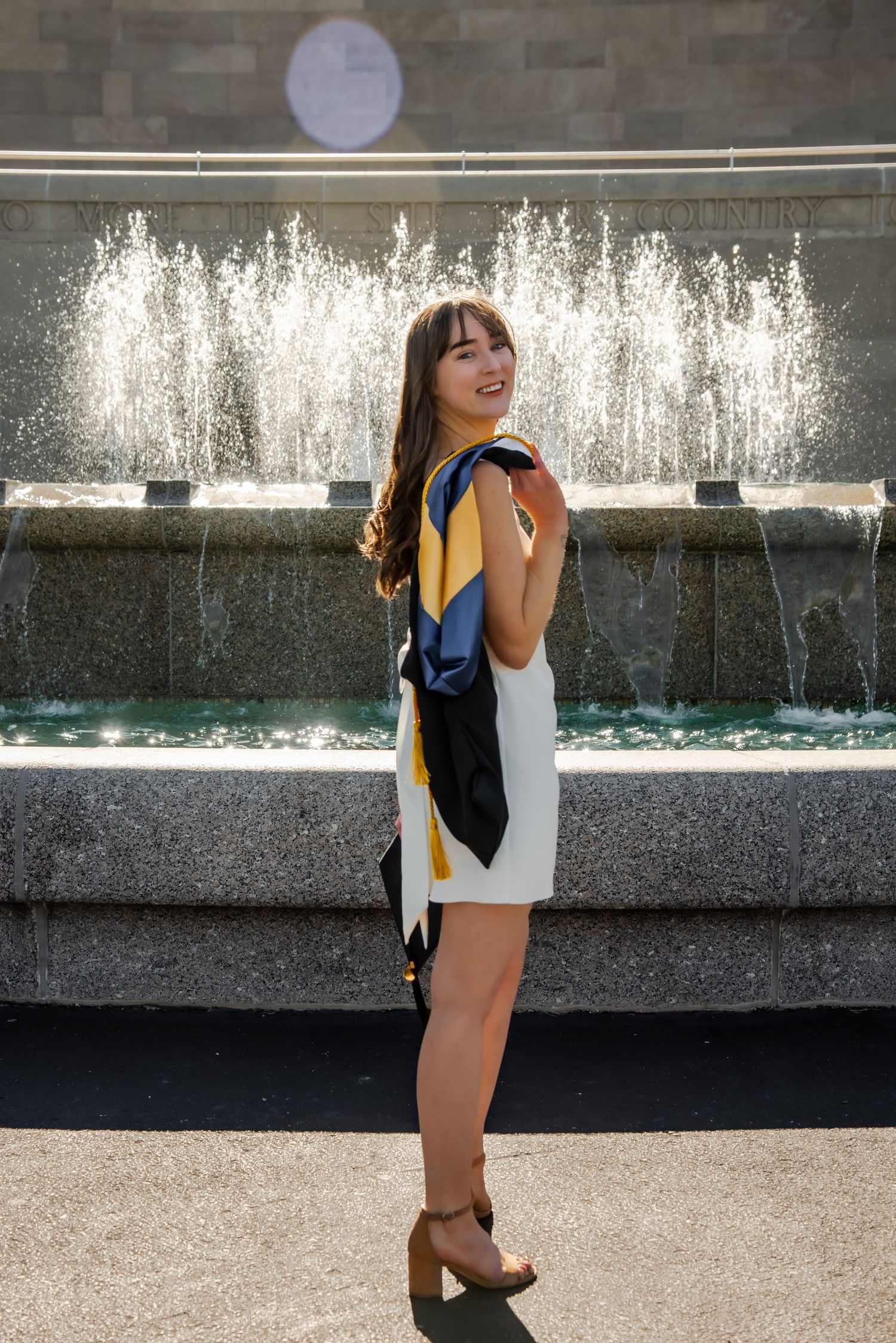 A person in a white dress and colorful scarf poses in front of a decorative water fountain on a sunny day.