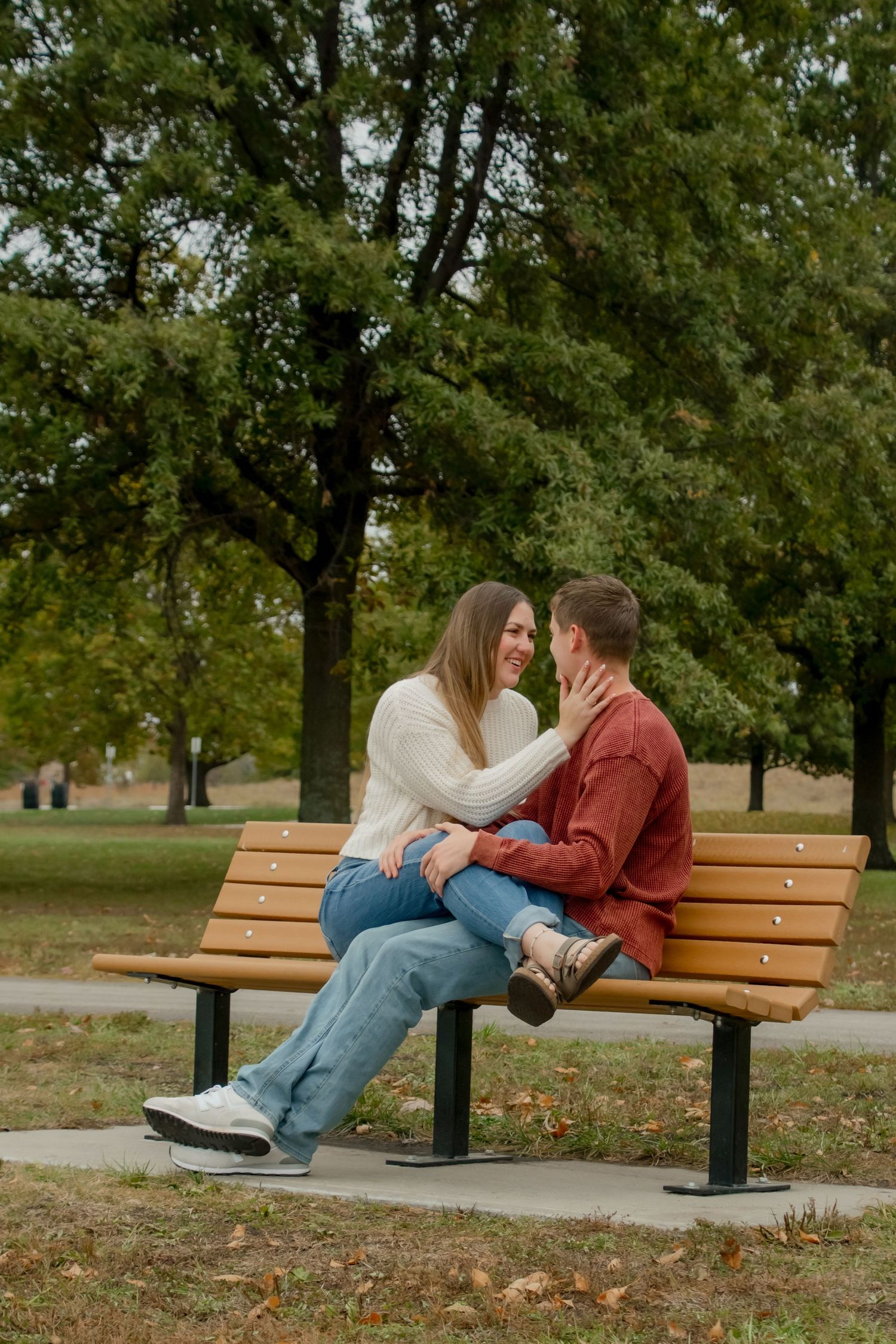 A couple shares a romantic moment sitting together on a wooden park bench surrounded by autumn trees.