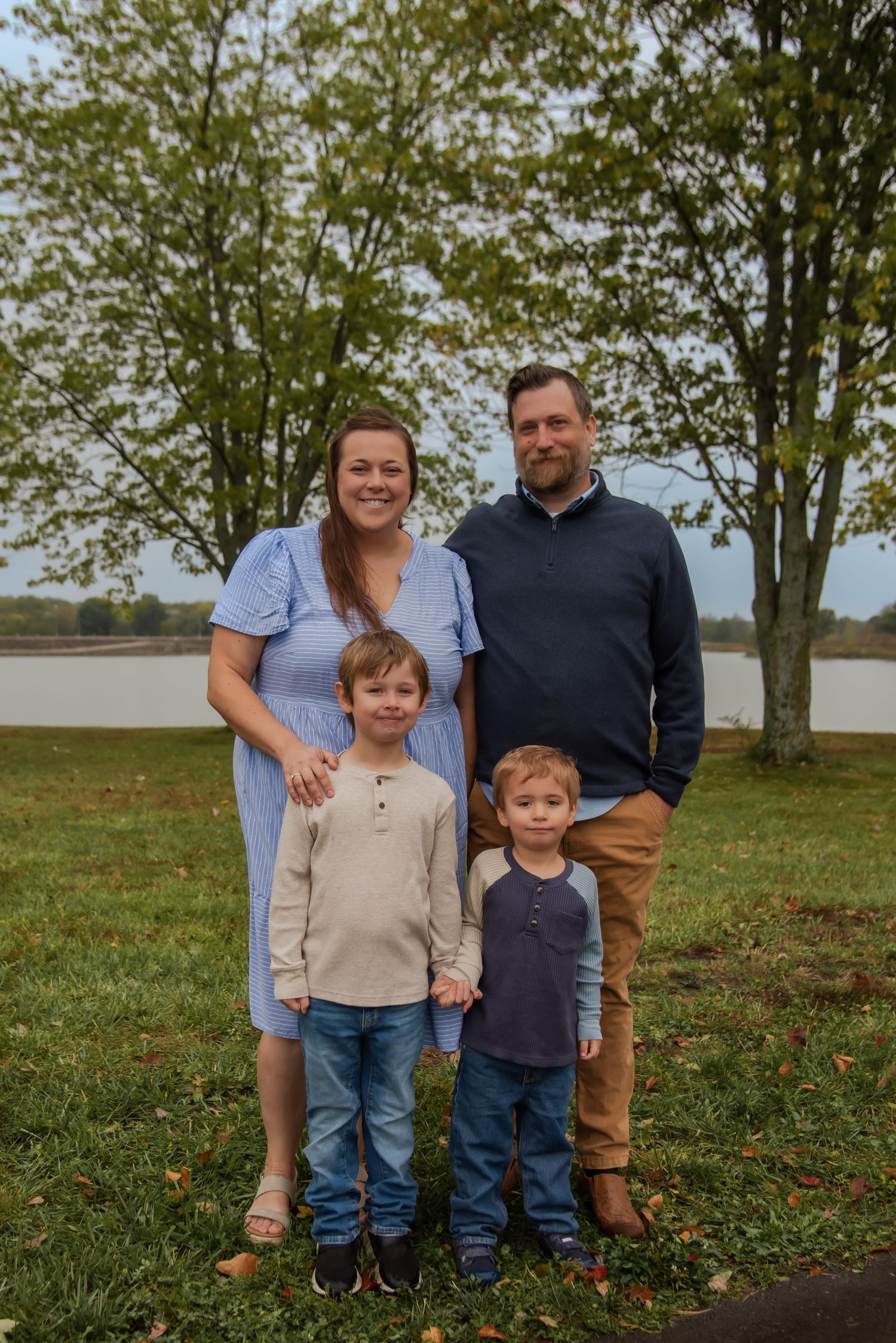 A family poses together outdoors on a fall day with trees and grass in the background.