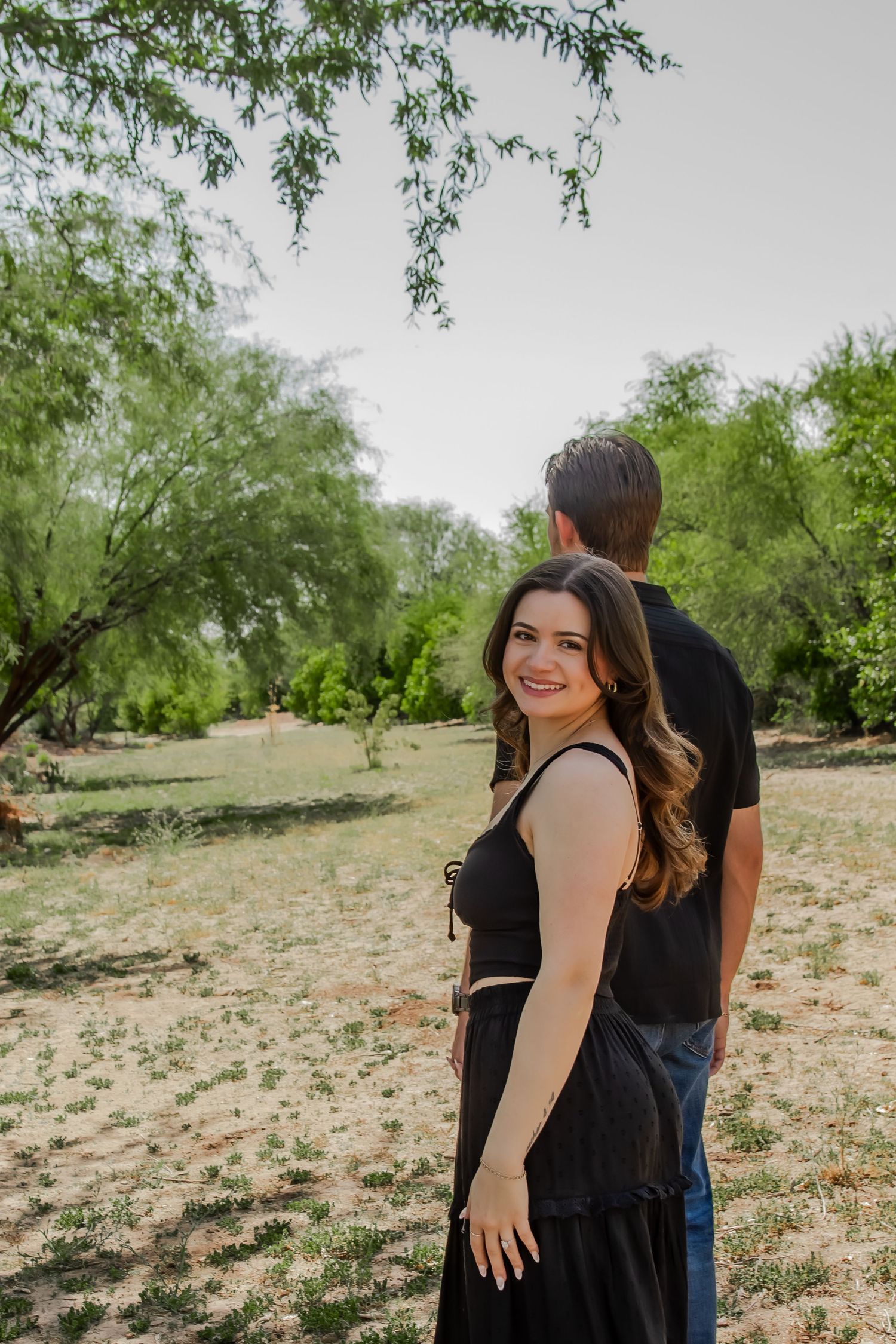 A couple in black clothing standing together in a desert landscape with mesquite trees on a sunny day.