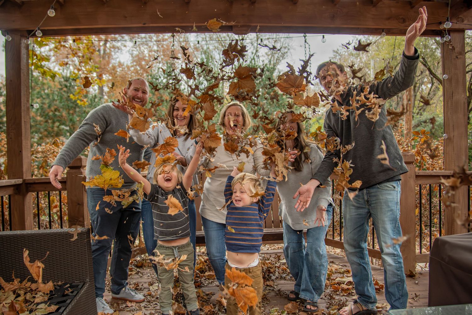 A group plays in fallen autumn leaves at a rustic wooden pavilion during a joyful outdoor photoshoot.