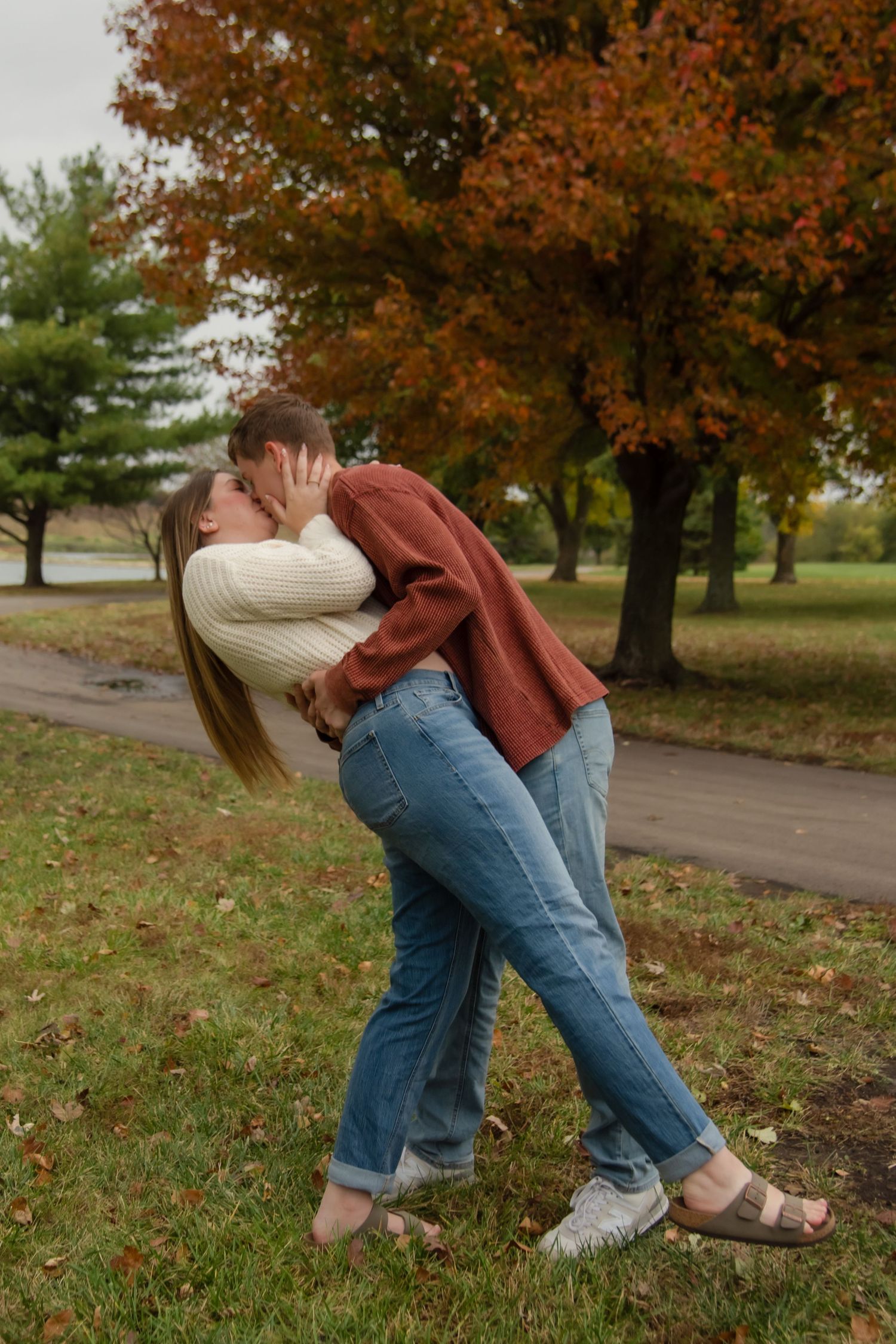 A couple in casual fall attire embraces playfully in a park with autumn foliage in the background.