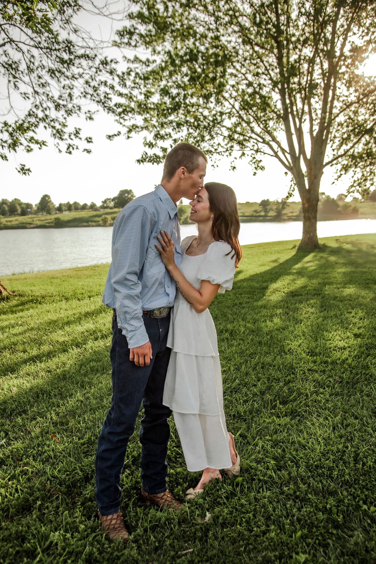 Two people share tender moments on a green lawn with trees and sunlight behind them.