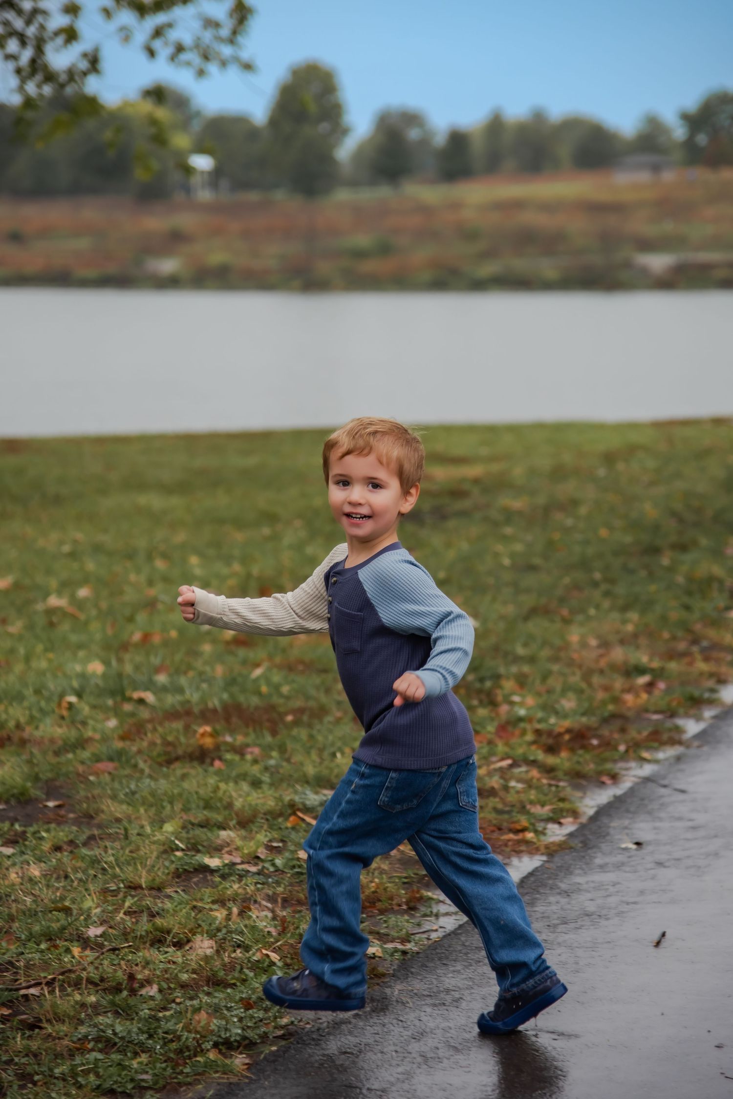 A small child wearing a navy vest and jeans walks playfully along a path near a lake on an autumn day.