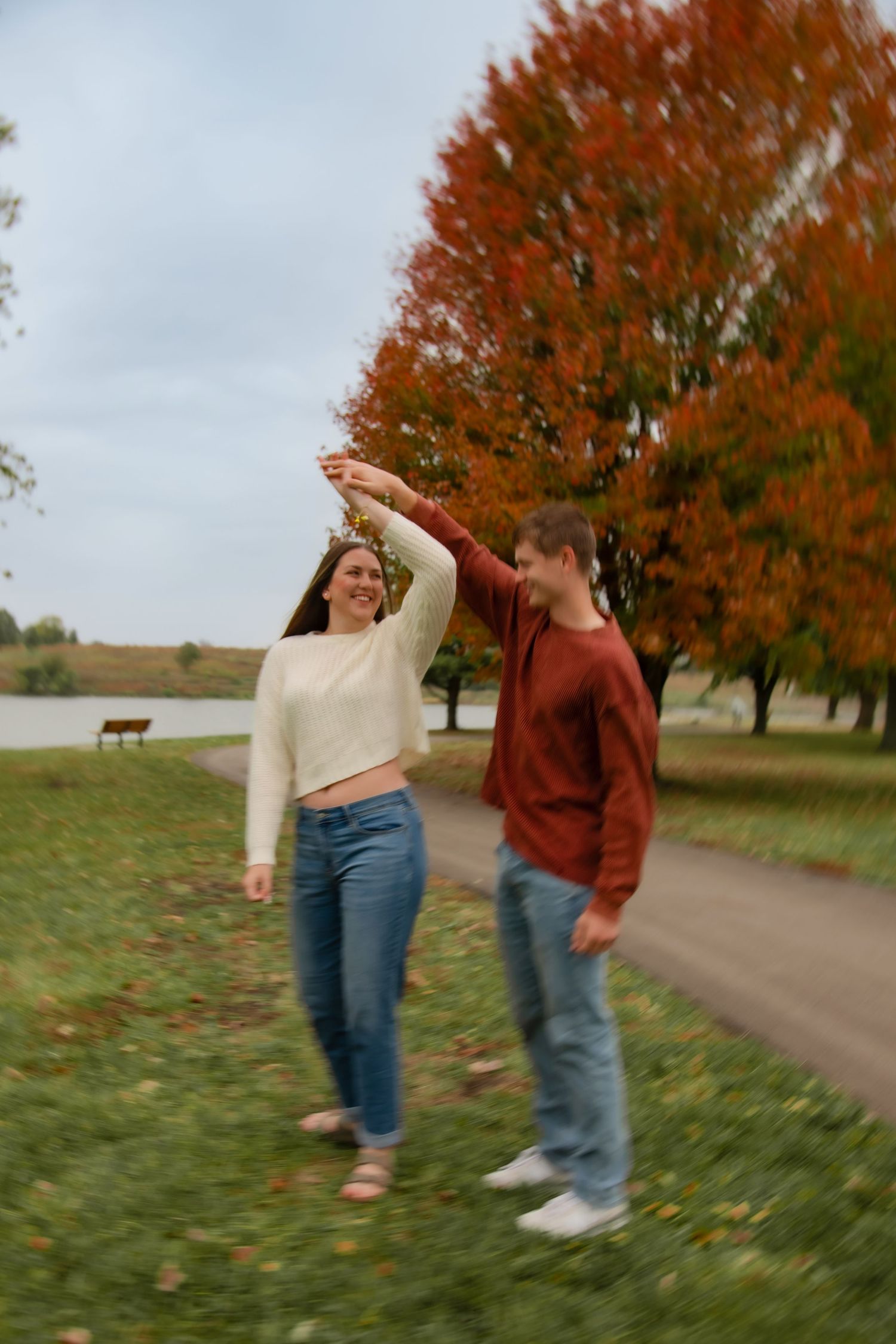 Two people dance and twirl together on a path with fall trees and orange leaves in the background.
