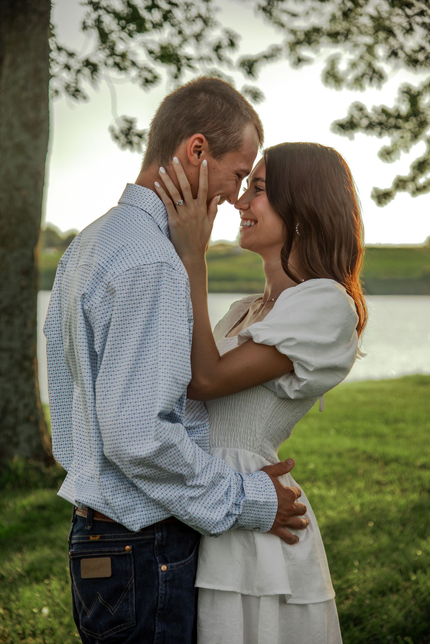 A romantic couple embraces near a lake during golden hour with trees in the background.