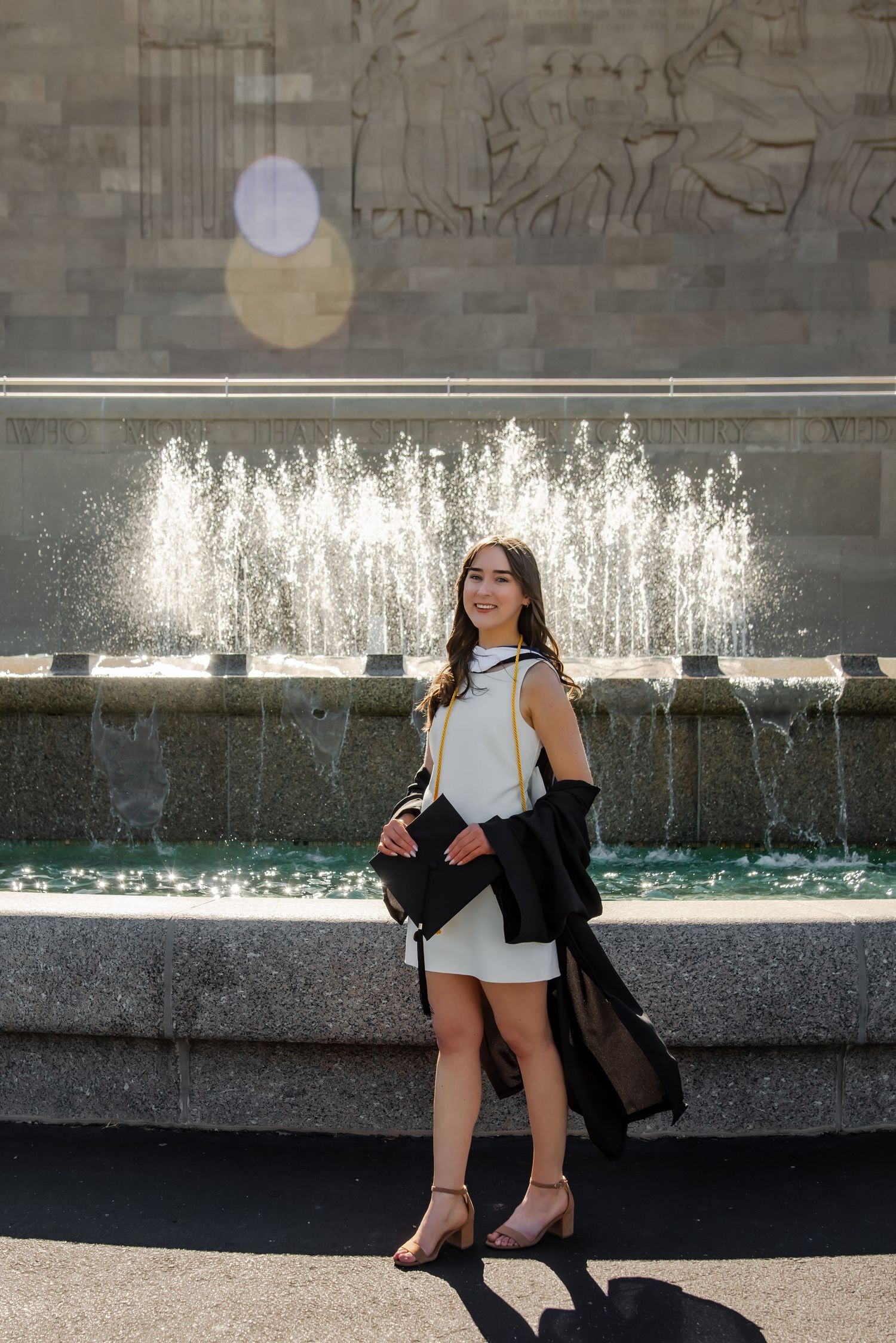 Graduate in black cap and gown sits by a fountain wearing a white dress.