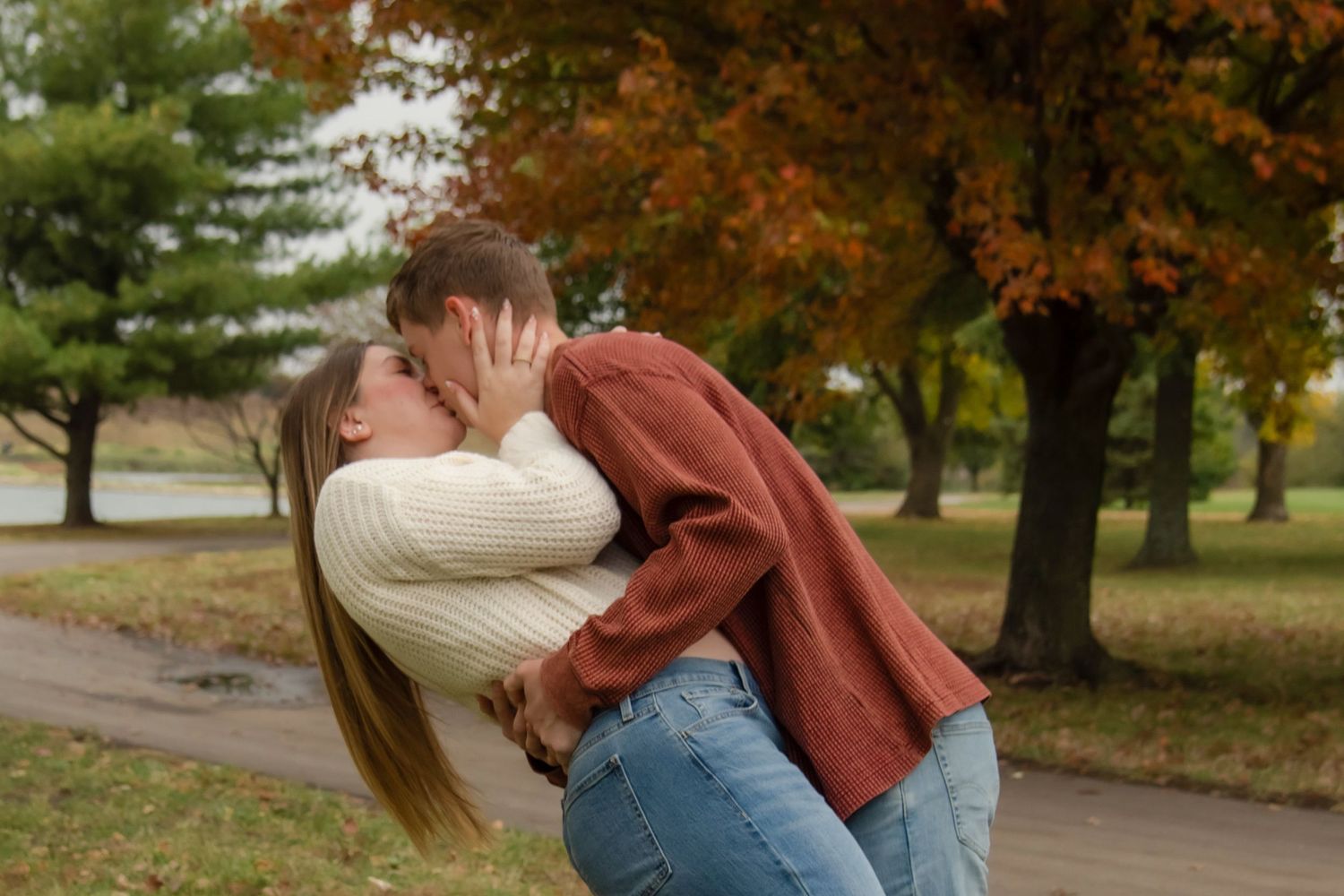 A couple shares a romantic embrace in a park with autumn foliage in the background.