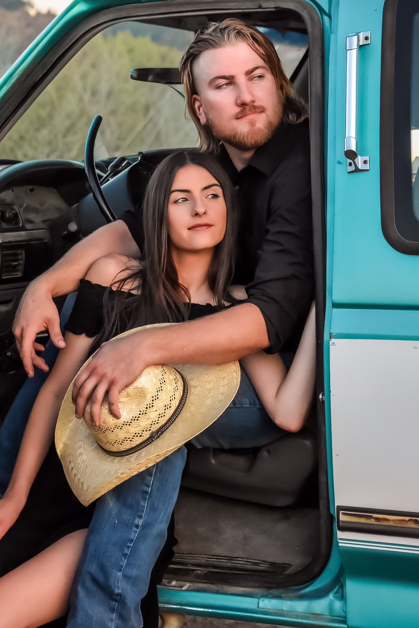 A romantic couple poses in a vintage turquoise truck with a straw hat for an engagement photo session.