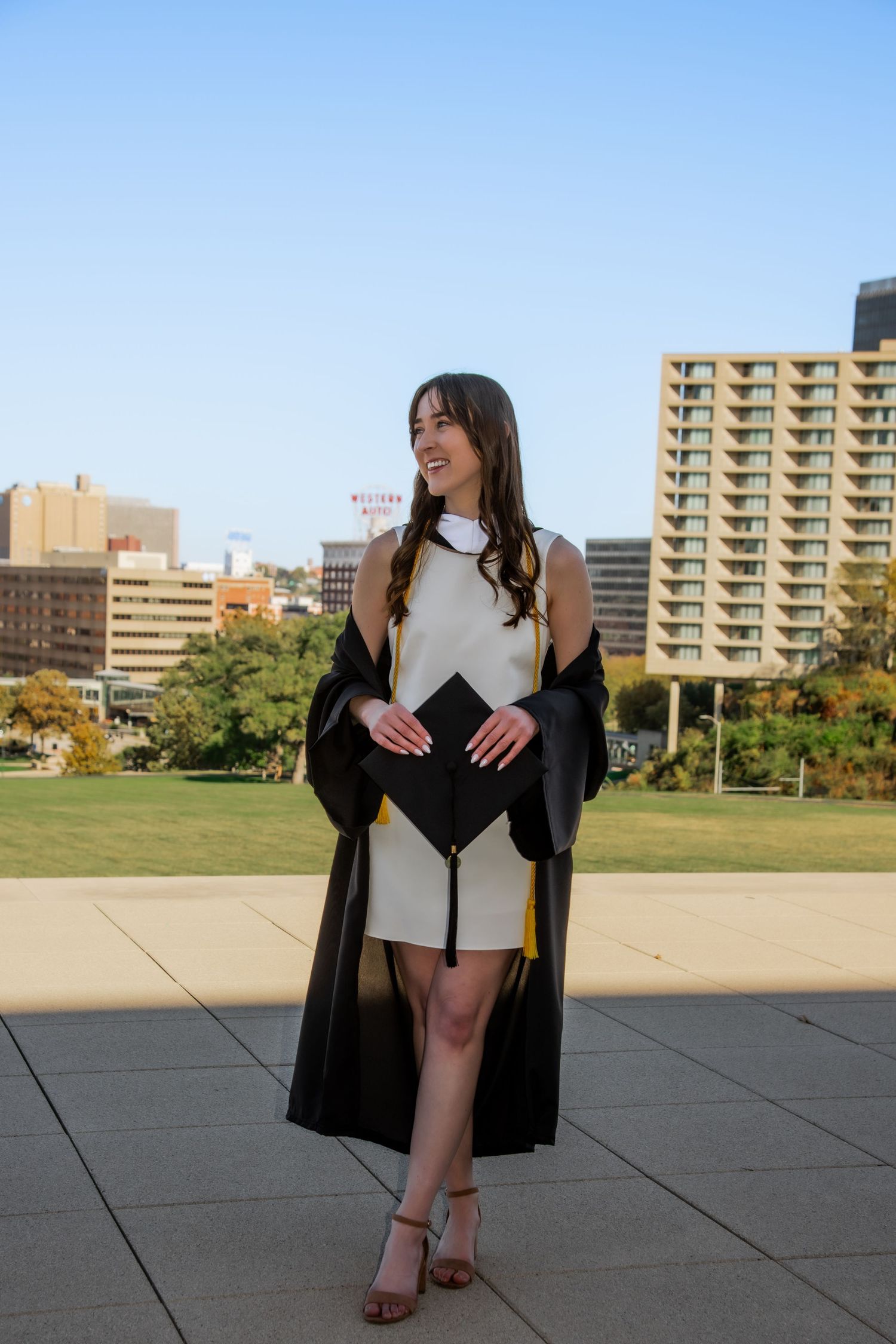 Graduate posing in black gown and white dress on campus grounds with buildings in background.