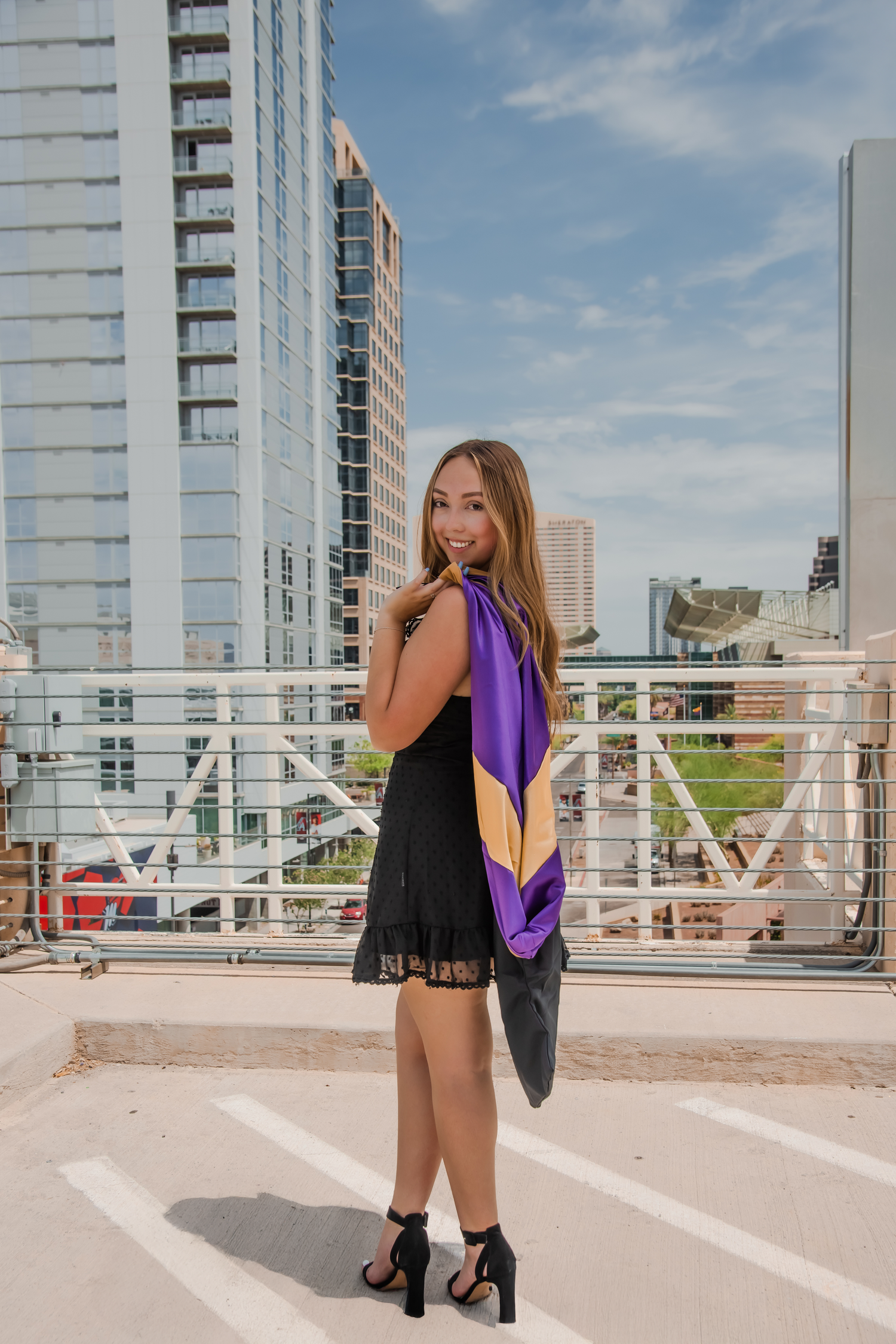 Person in purple top and black dress poses on urban rooftop with city buildings in background.