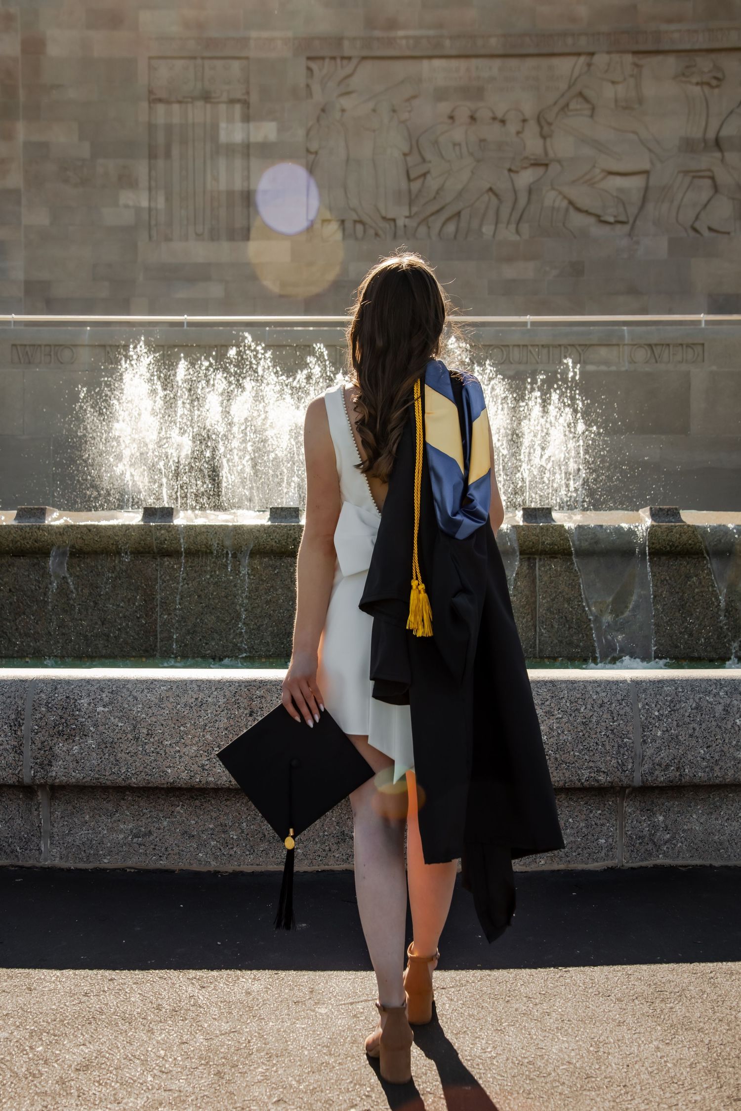 Two graduates in black and white attire walk past a fountain wall during a graduation ceremony.