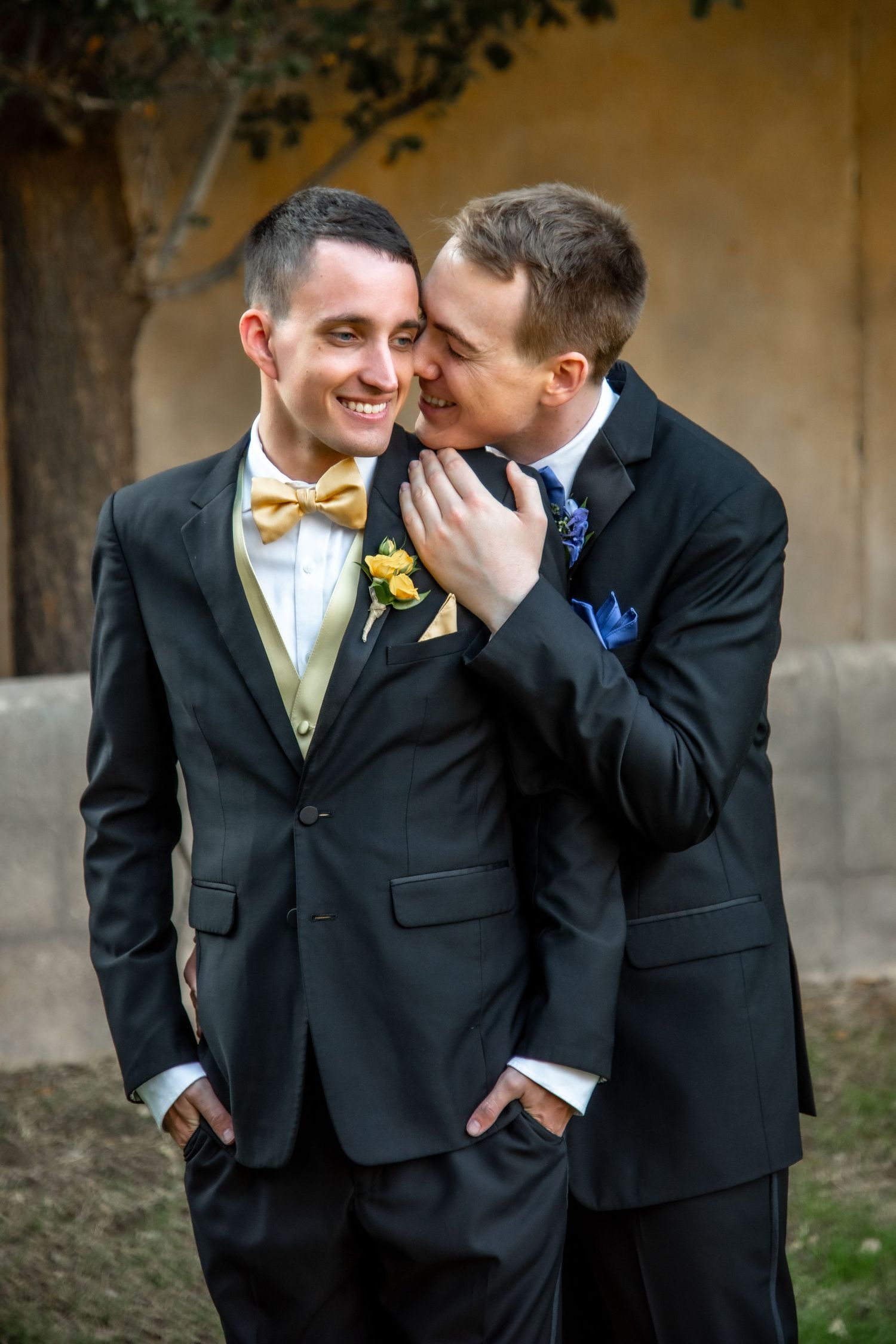 Two people in black suits share an intimate moment at a wedding, one wearing a gold bowtie and yellow boutonniere.