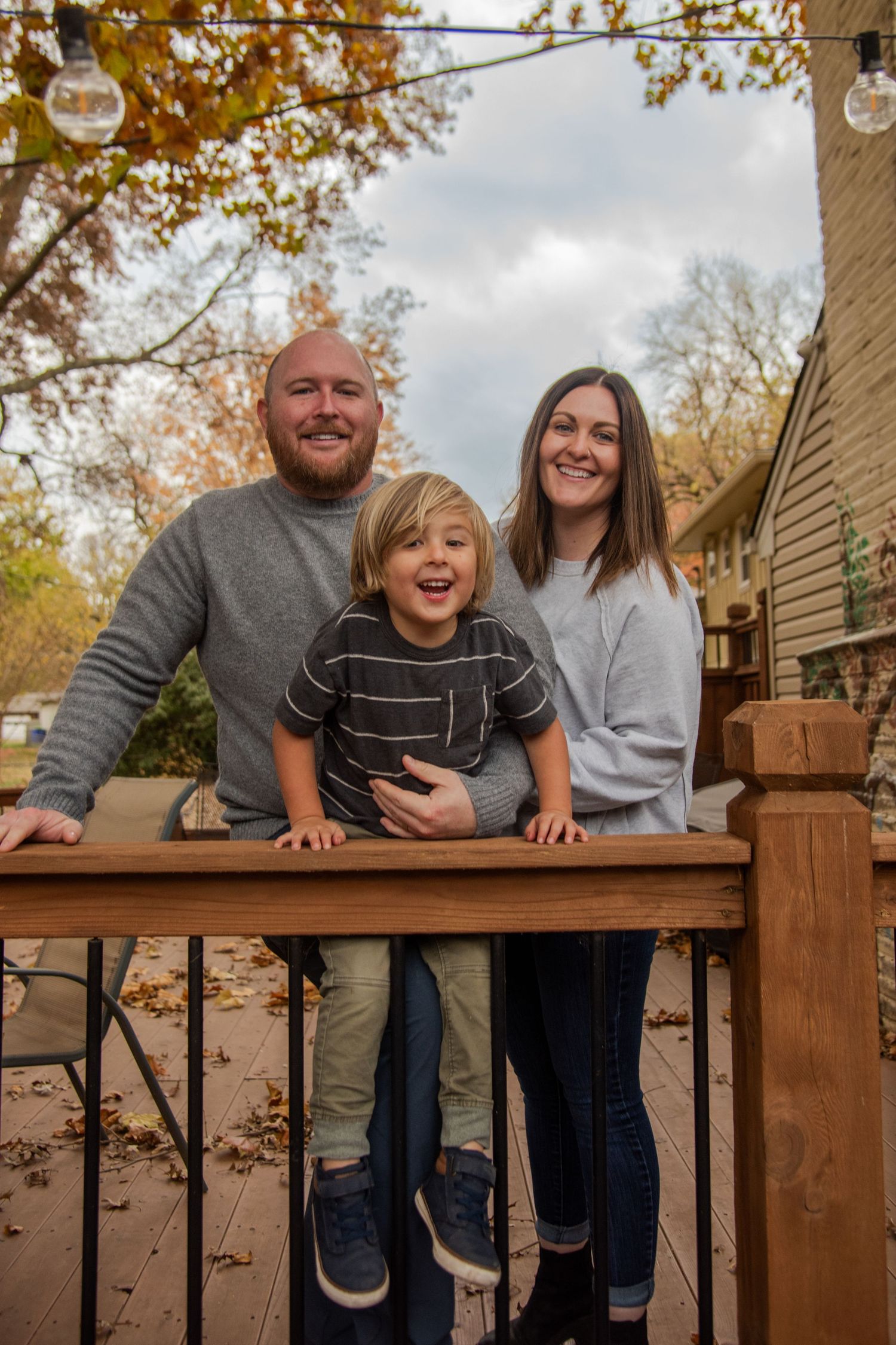 Family members lean on a wooden deck railing during an autumn photoshoot with fall foliage in background.