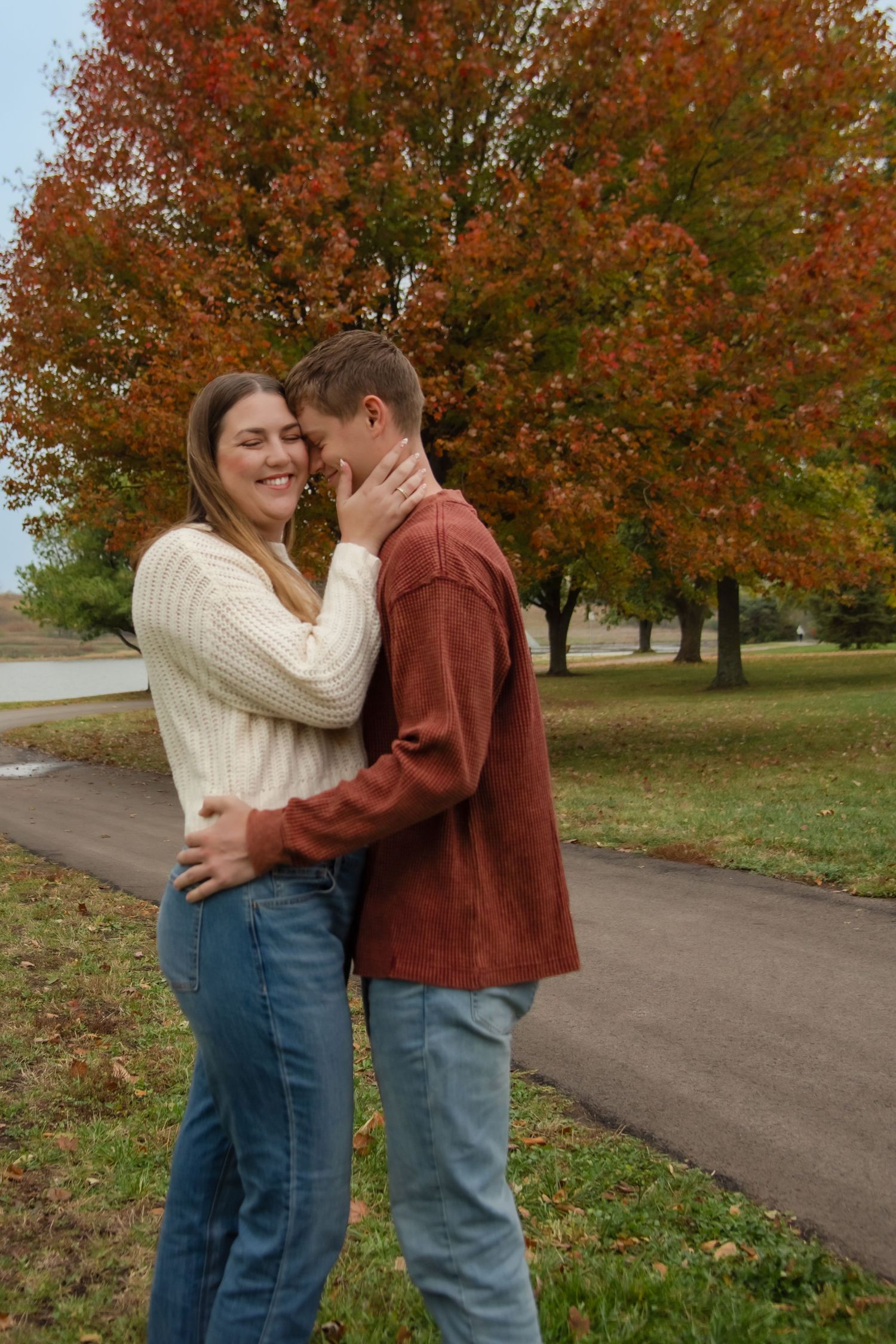 A romantic couple embraces on a pathway surrounded by autumn trees with vibrant orange foliage.